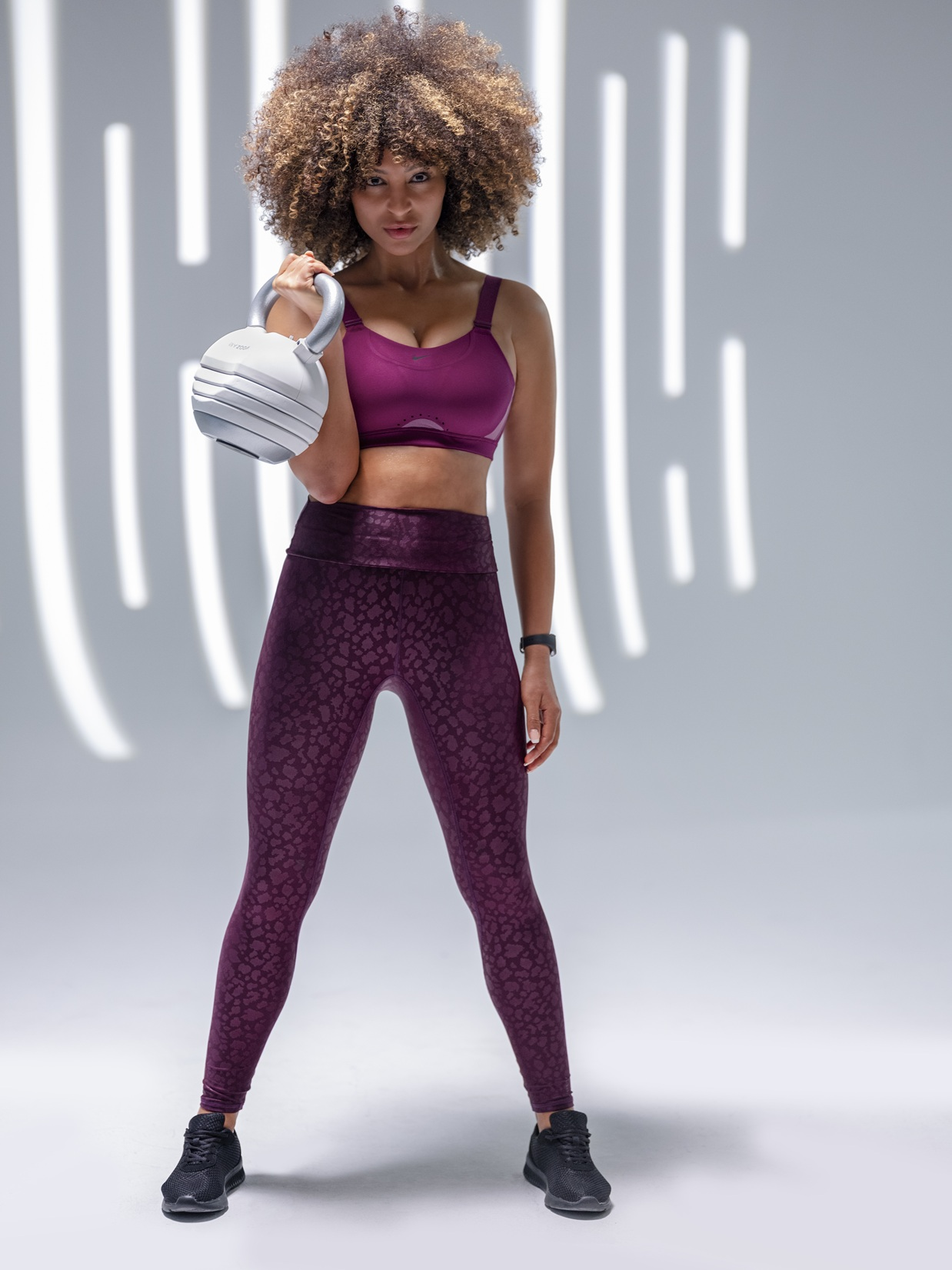 Frau mit lockigem Haar hält eine weiße Kettlebell, trägt lila Sportkleidung.