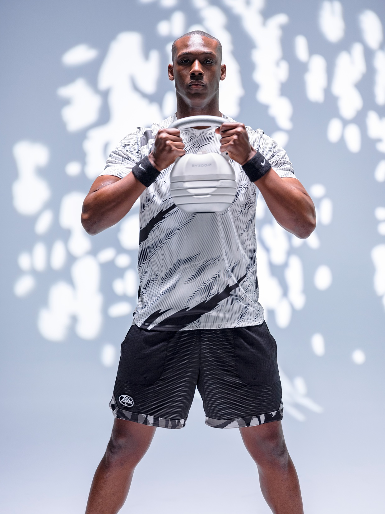 Ein Mann in Sportkleidung hält ein weißes Kettlebell vor einem hellen Hintergrund mit Schatten.