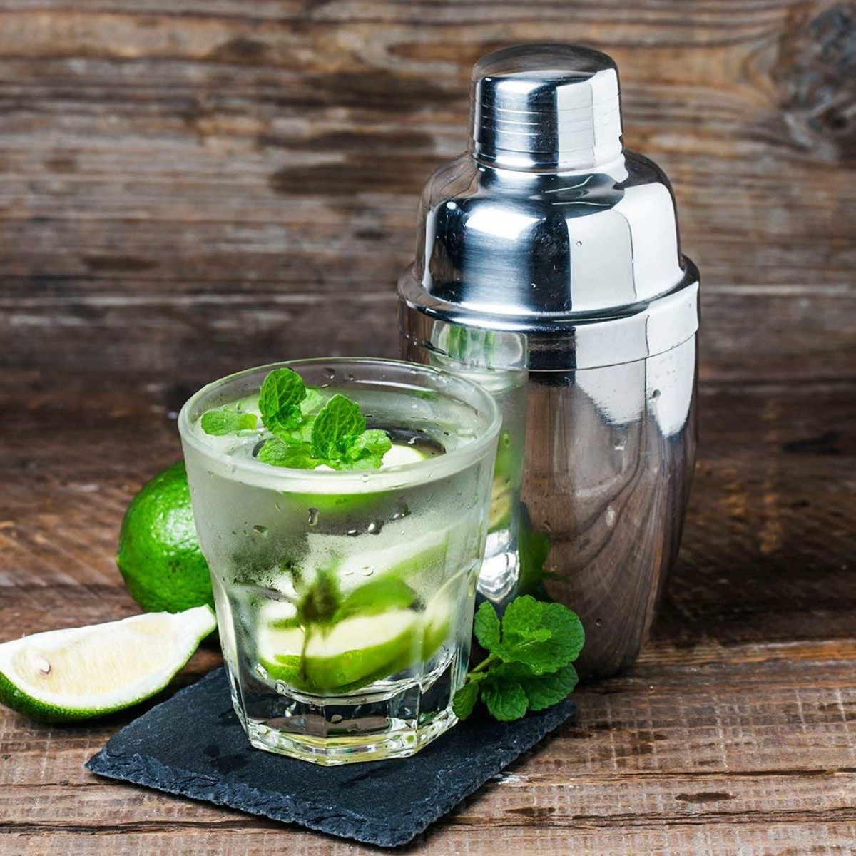 Ein Glas mit einem Getränk, Minze und Limette, daneben ein Shaker.