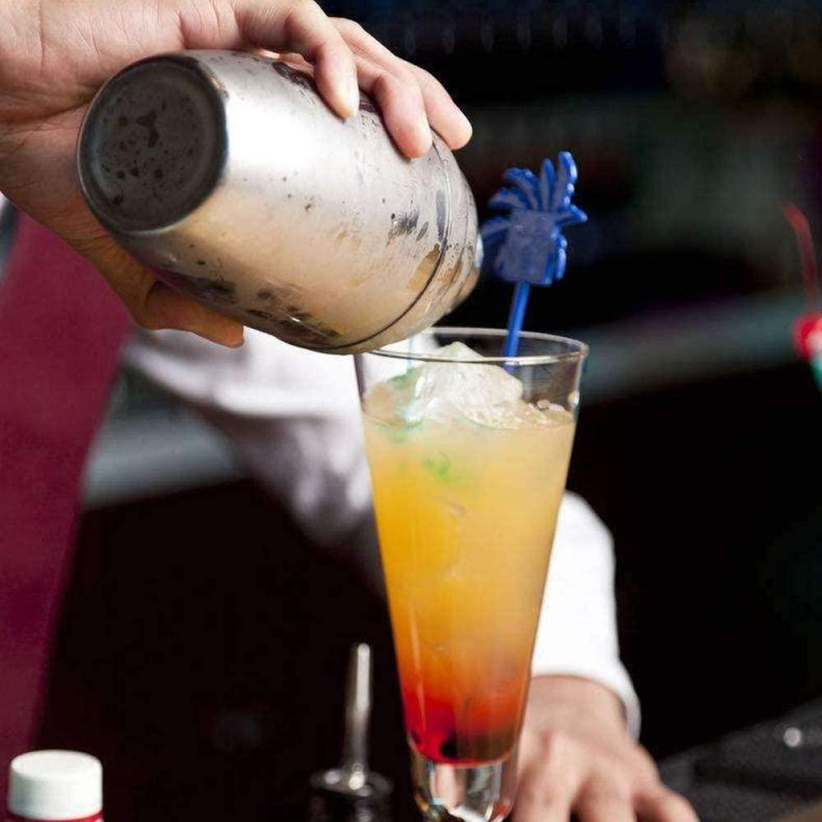 Ein Barkeeper gießt einen bunten Cocktail aus einem Metallshaker in ein Glas mit Eis.