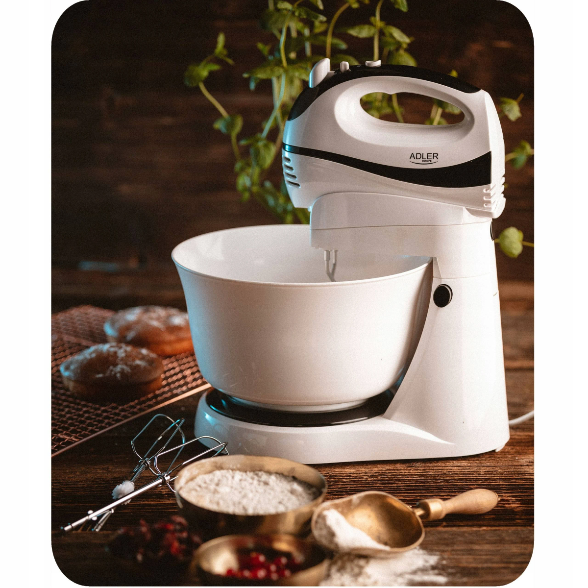 Biały mikser ręczny z misą, składnikami i ciastkami na drewnianym stole.