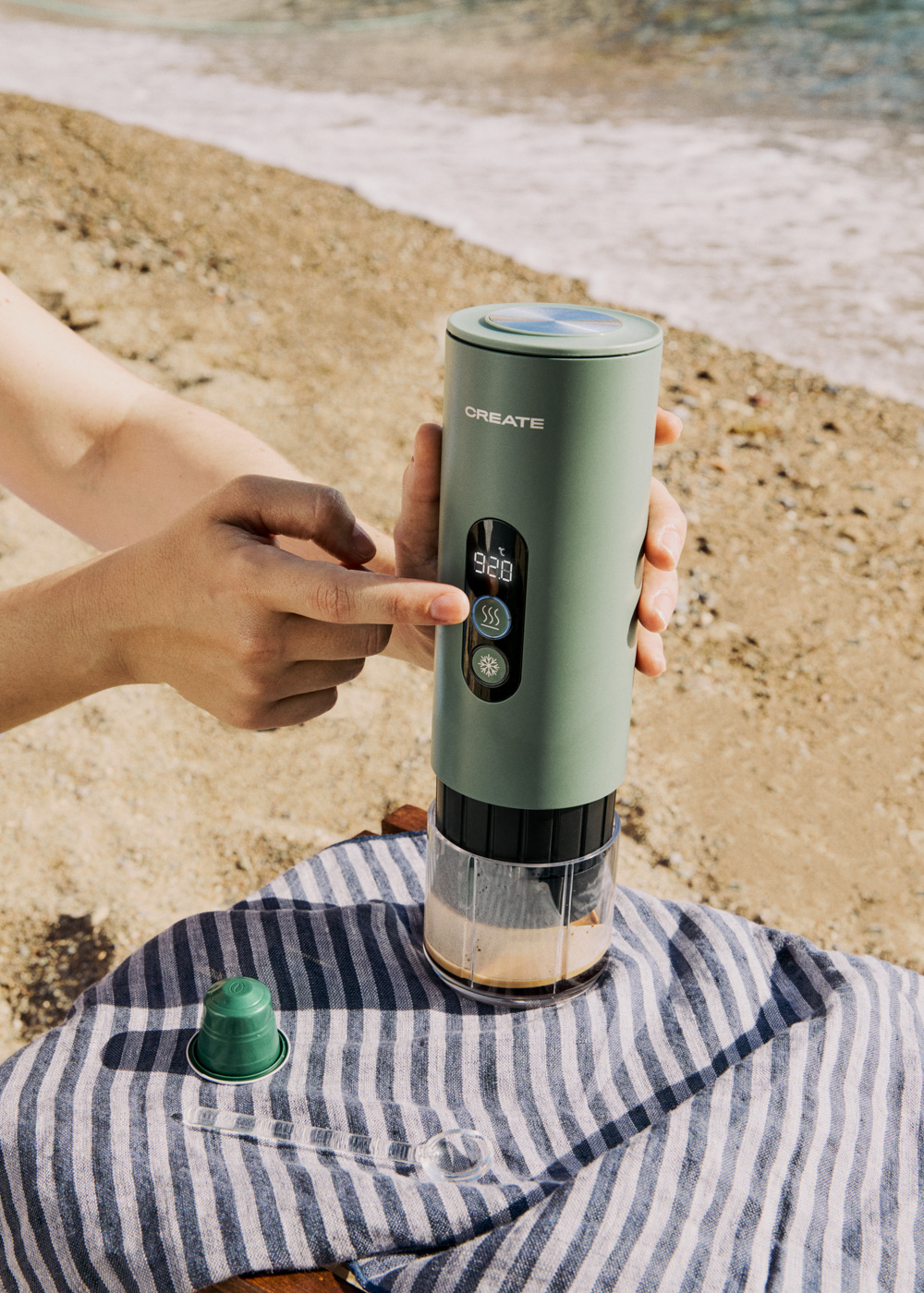 Cafetera verde en la playa, manos operándola.