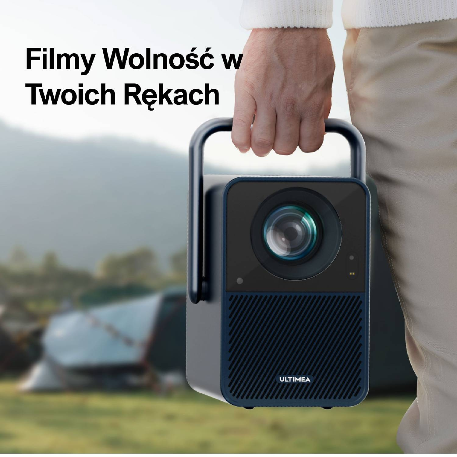 Osoba trzyma przenośny projektor. W tle rozstawione są namioty.