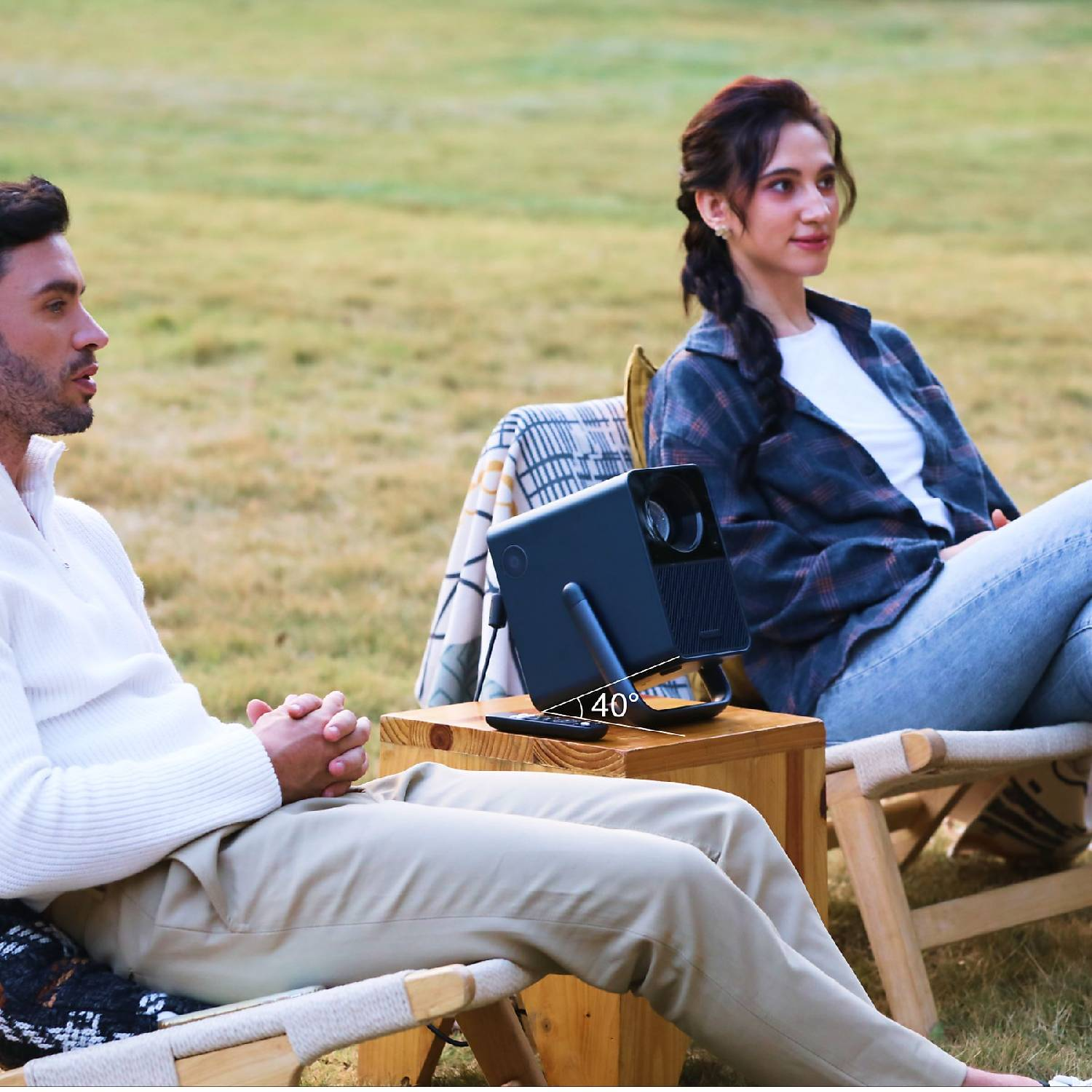 Mężczyzna i kobieta siedzą na zewnątrz na krzesłach, patrząc w prawo. Projektor stoi na drewnianym stole.