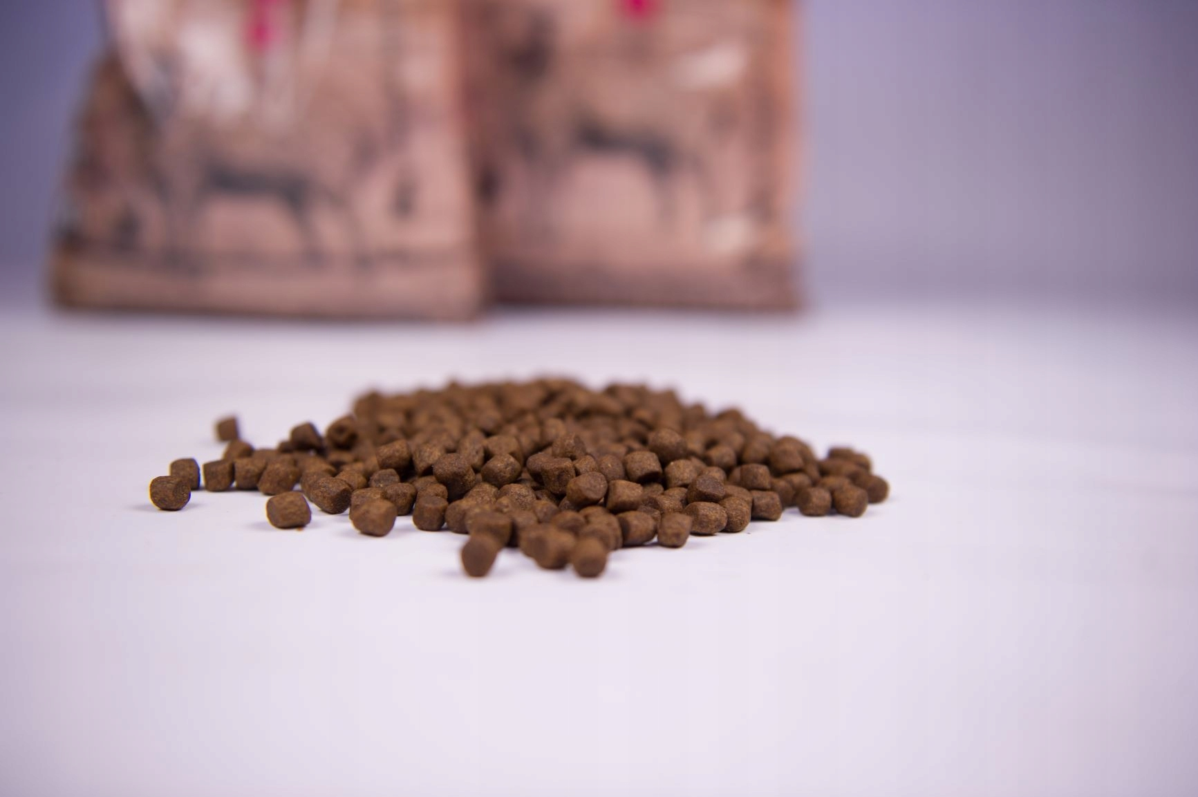 Stos brązowej karmy na białej powierzchni, z niewyraźnymi workami w tle.