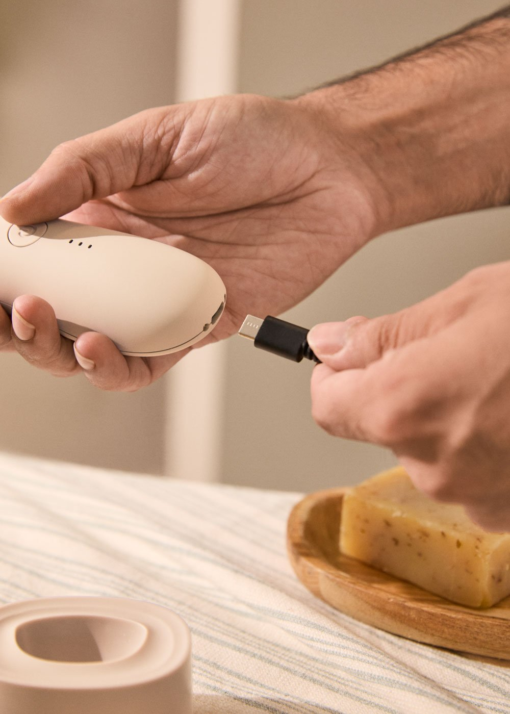 Hombre enchufa cable USB-C a dispositivo beige. Jabón en plato madera.