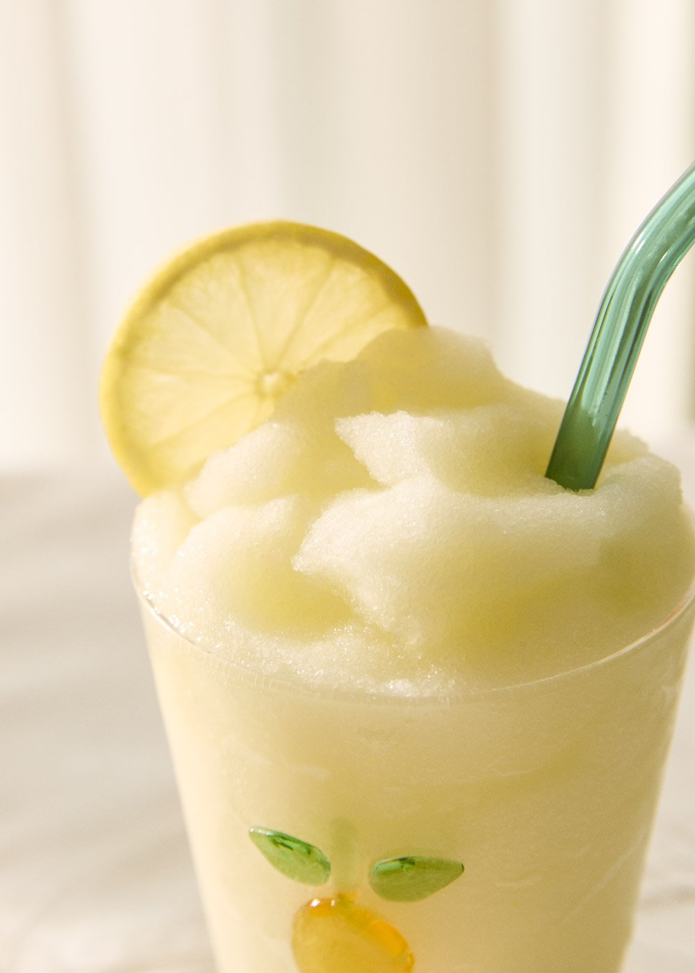 Una bebida de limón congelada en un vaso con una rodaja de limón y una pajita verde.