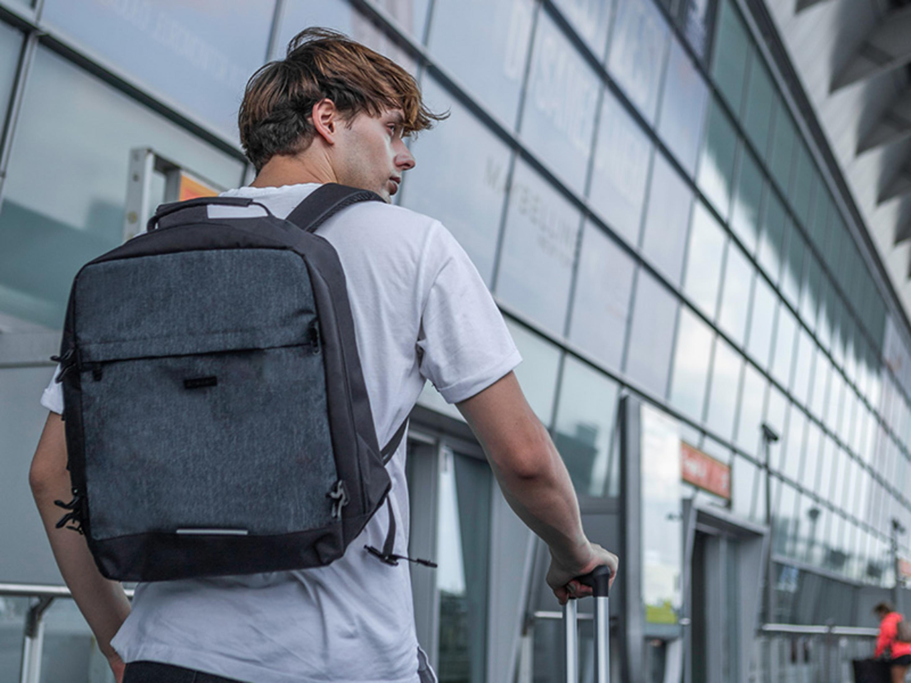 Un uomo con uno zaino tira una valigia davanti a una facciata in vetro.