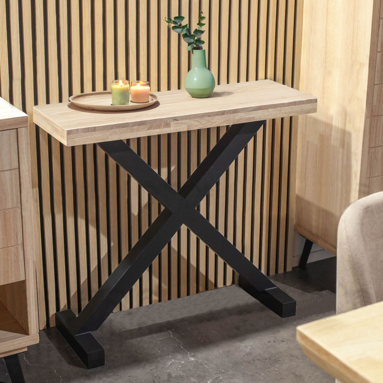 Una mesa estrecha de madera con estructura en X negra, velas y jarrón con ramas.