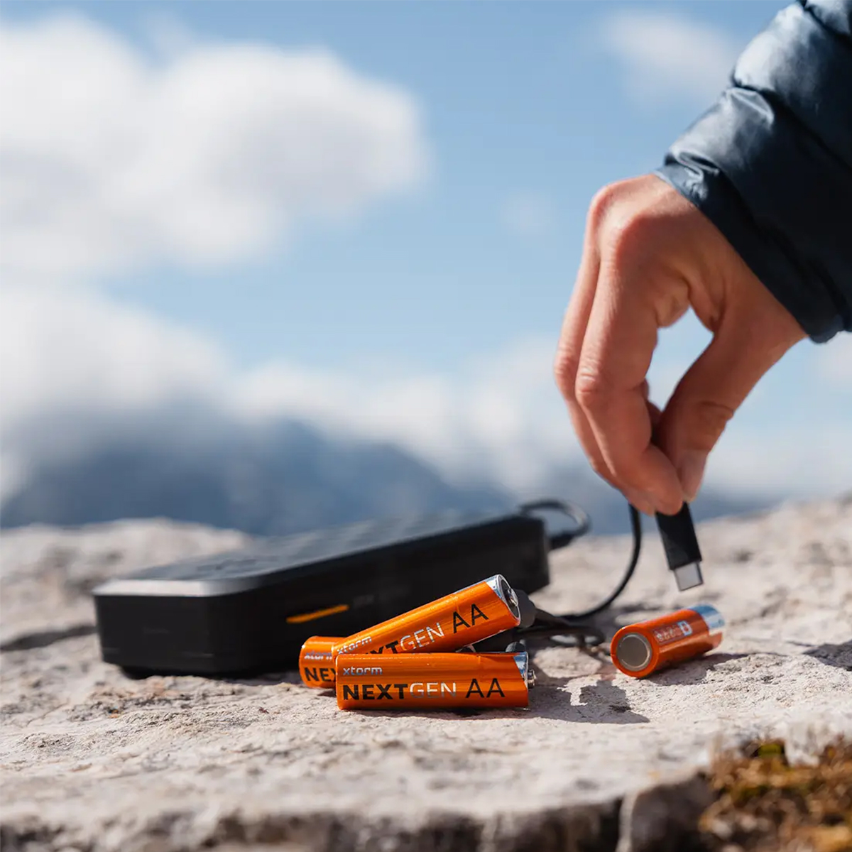 Mano che tiene cavo di ricarica vicino a batterie arancioni e power bank nero.