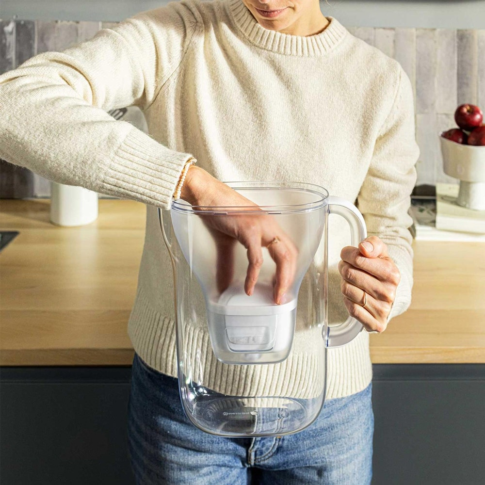 Una persona con maglione crema e jeans inserisce un filtro in una caraffa d'acqua trasparente.
