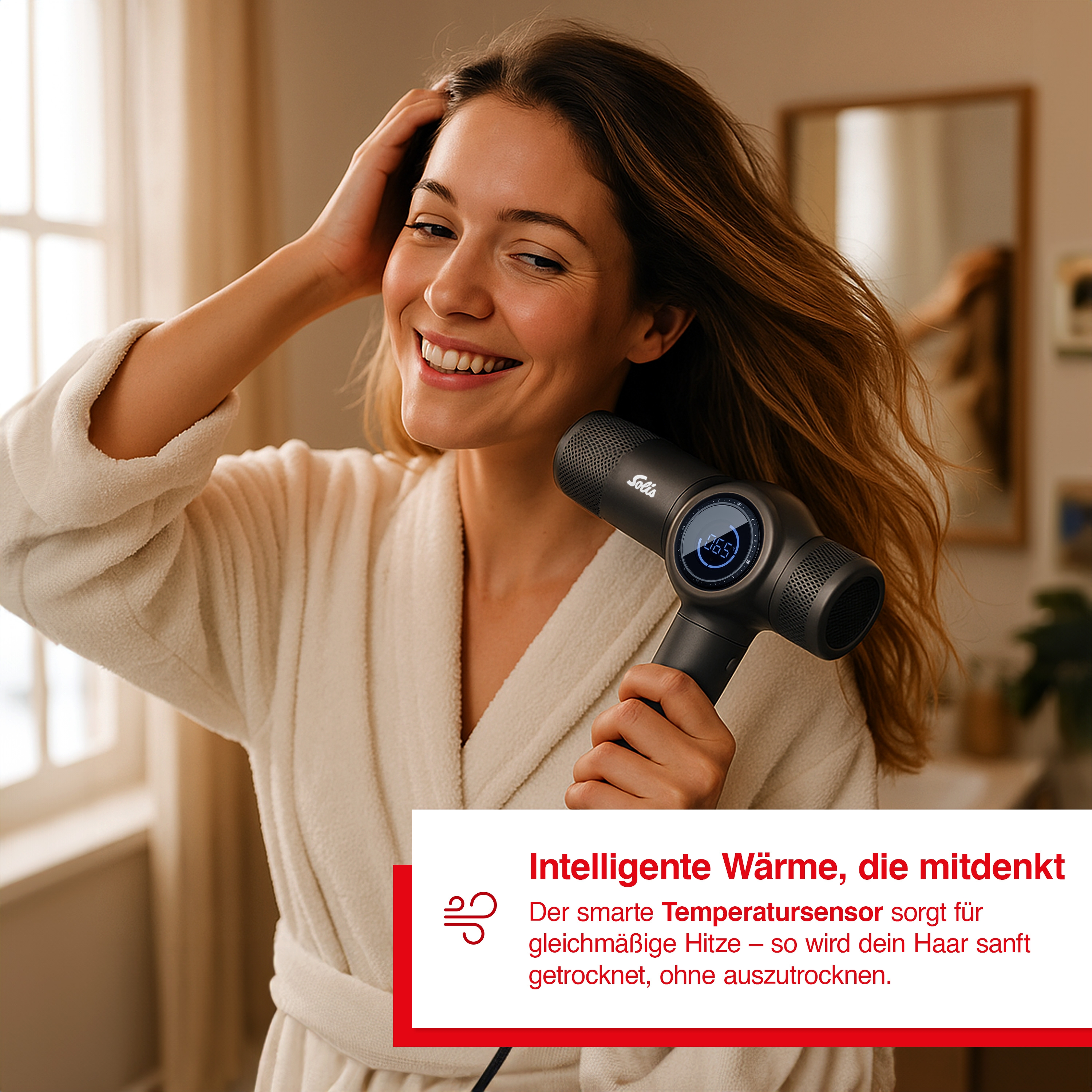 Eine Frau im Bademantel hält einen schwarzen Haartrockner, lächelt und fasst sich ans Haar.