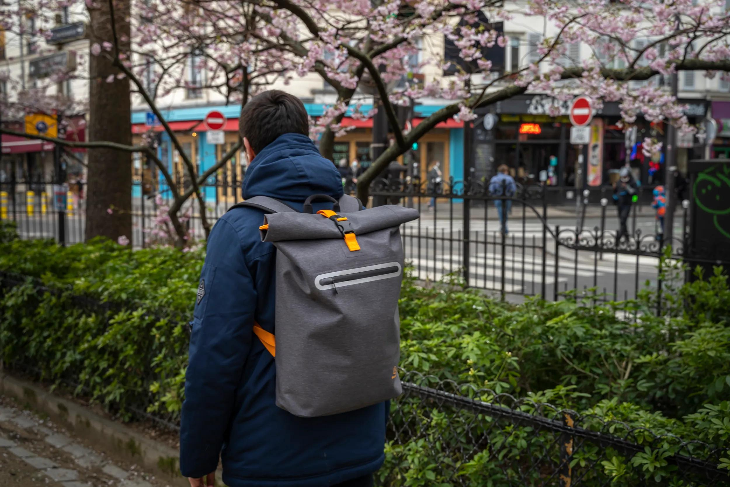 Mann mit grauem Rucksack und blauer Jacke geht an blühendem Baum vorbei.