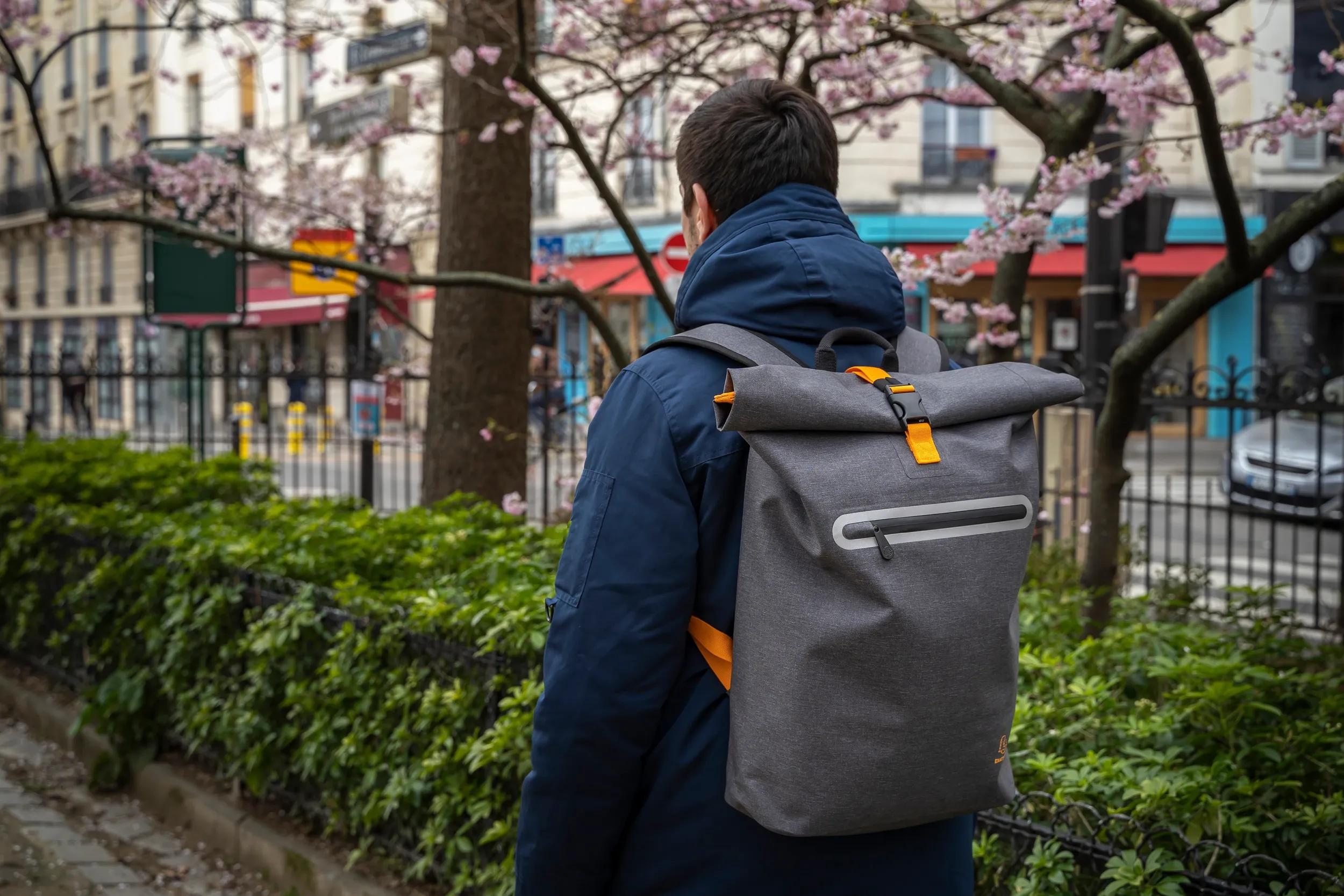 Mann mit grauem Rucksack in dunkler Jacke, blühende Bäume im Hintergrund.