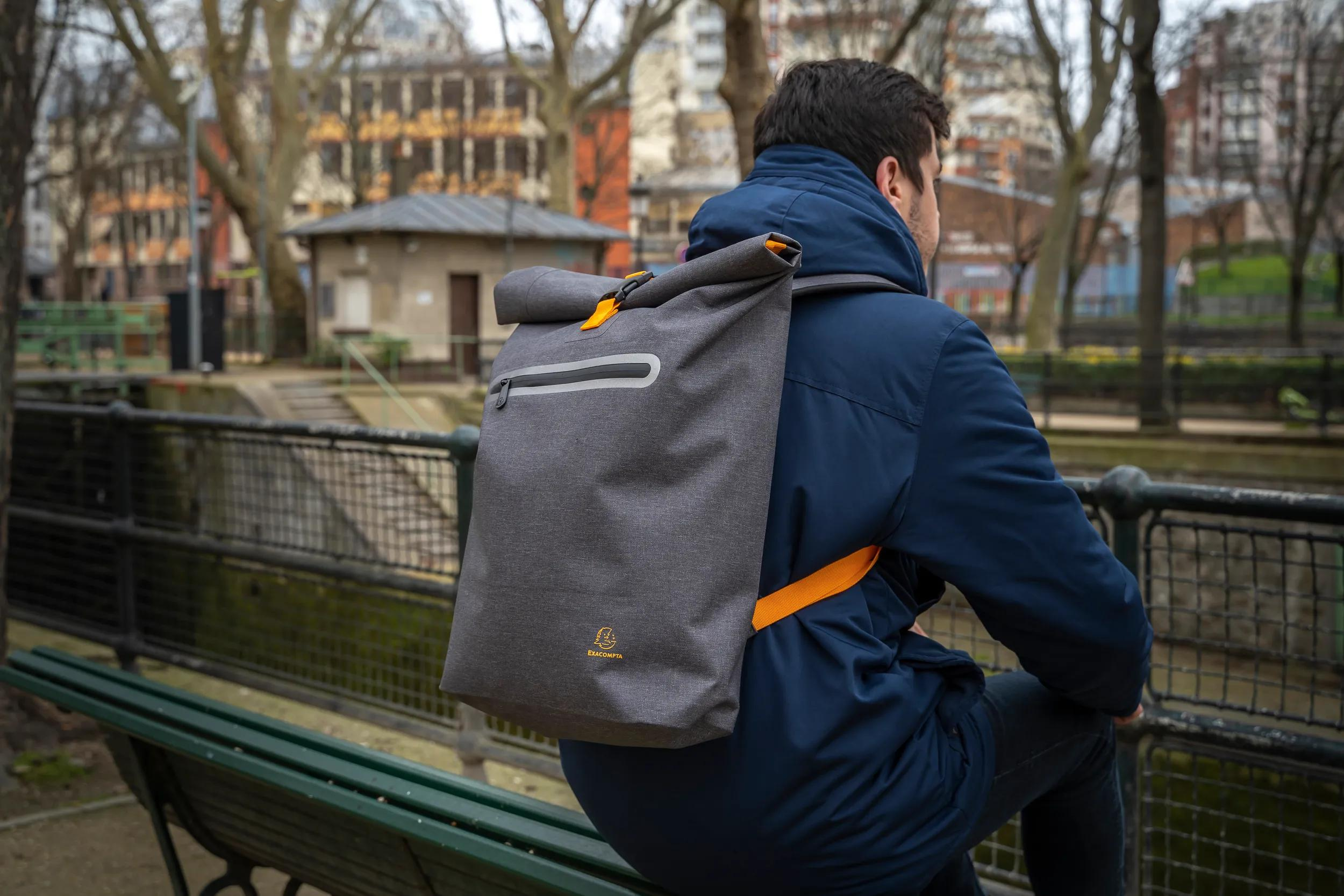 Mann mit grauem Rucksack sitzt auf einer Parkbank, Bäume und Gebäude im Hintergrund.