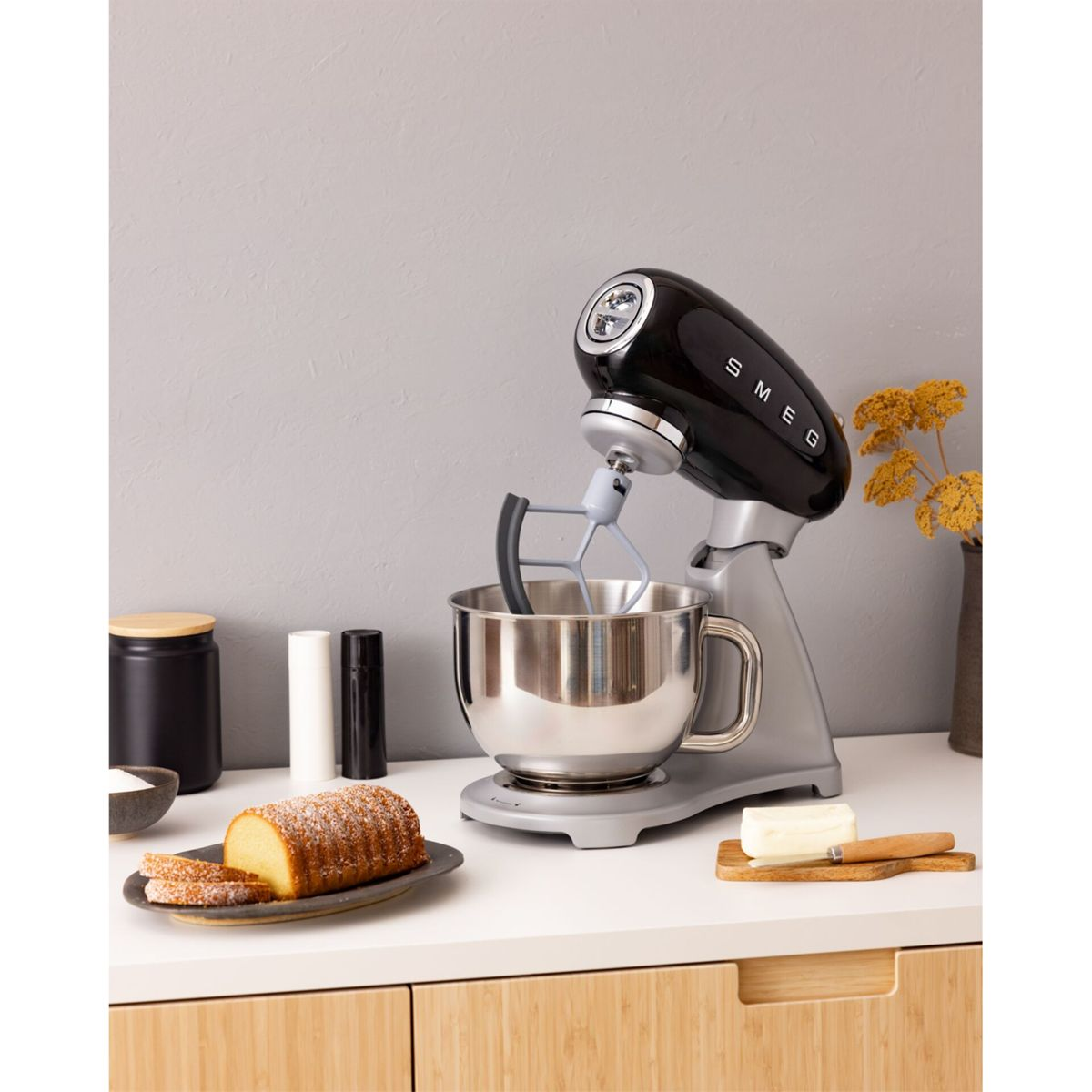 Gümüş kaseye sahip siyah Smeg stand mikseri, yanında kek ve tereyağı.