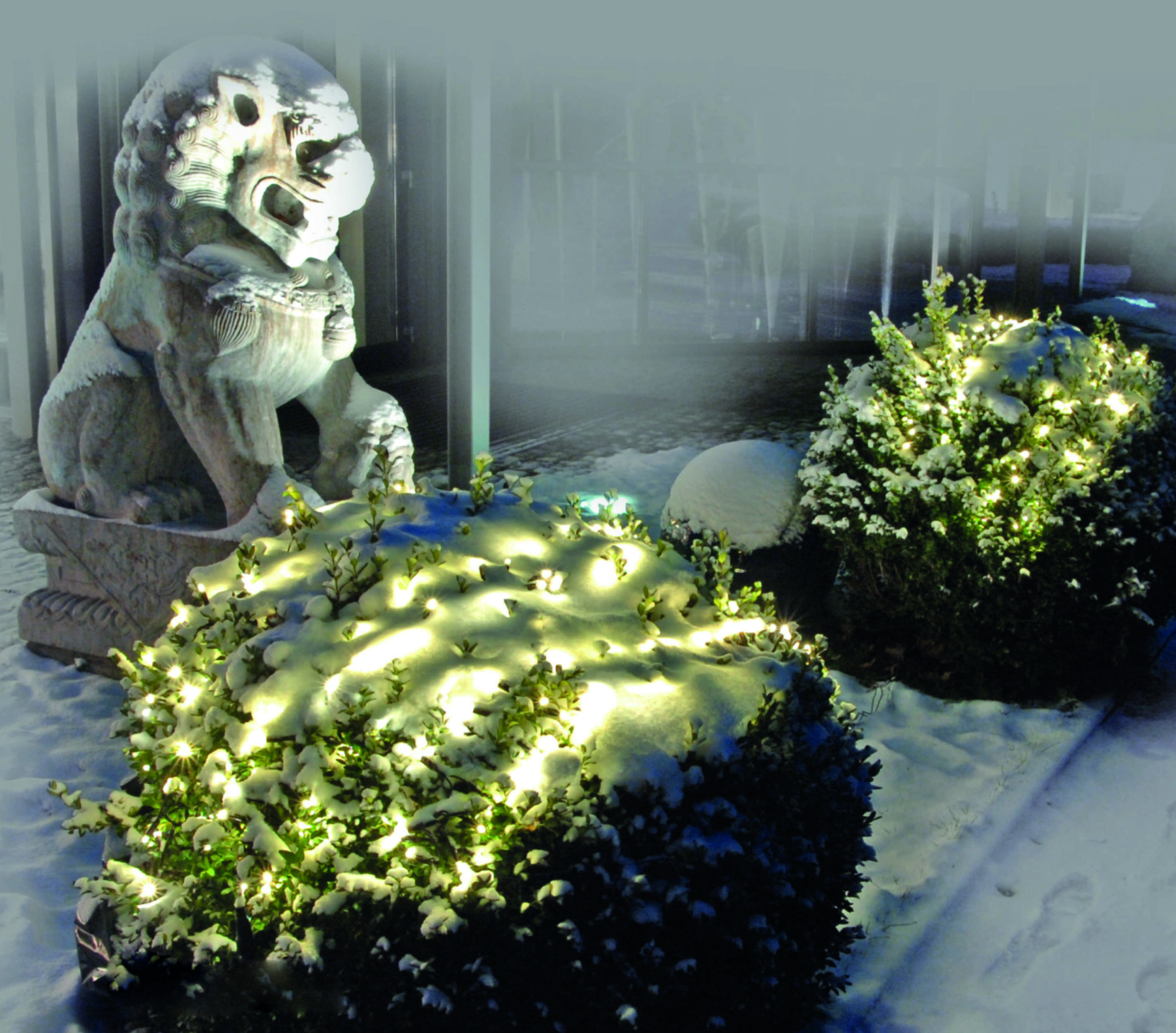 Ein steinerner Löwe steht im Schnee neben beleuchteten Büschen.