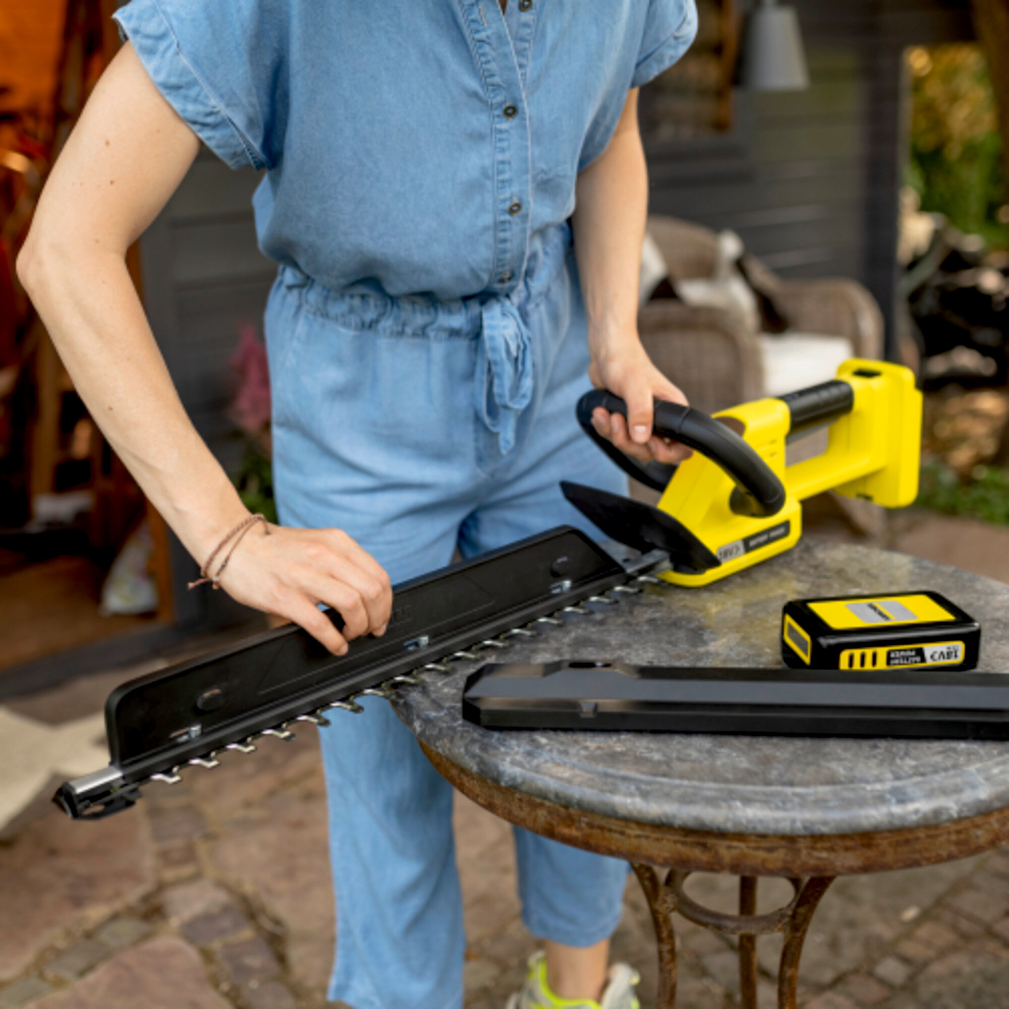 Frau im Jeans-Overall mit Akku-Heckenschere und Ersatzklinge auf einem Tisch.