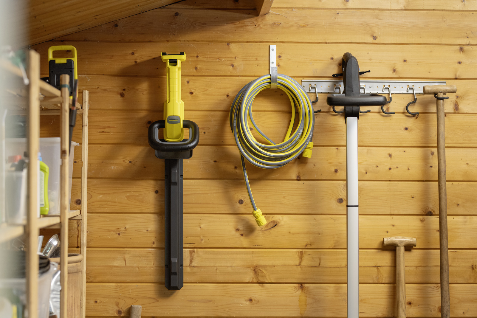 Gartengeräte an Holzwand: Heckenschere, Schlauch, Trimmer, Rechen.