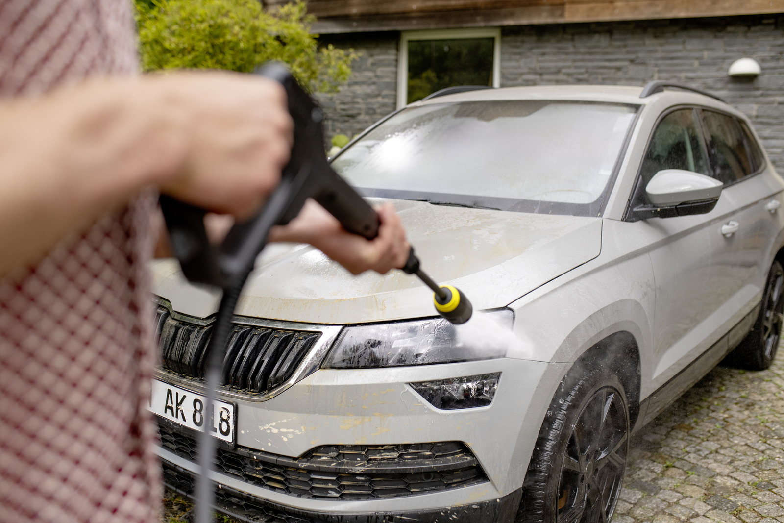Osoba myje szary samochód myjką ciśnieniową przed domem.