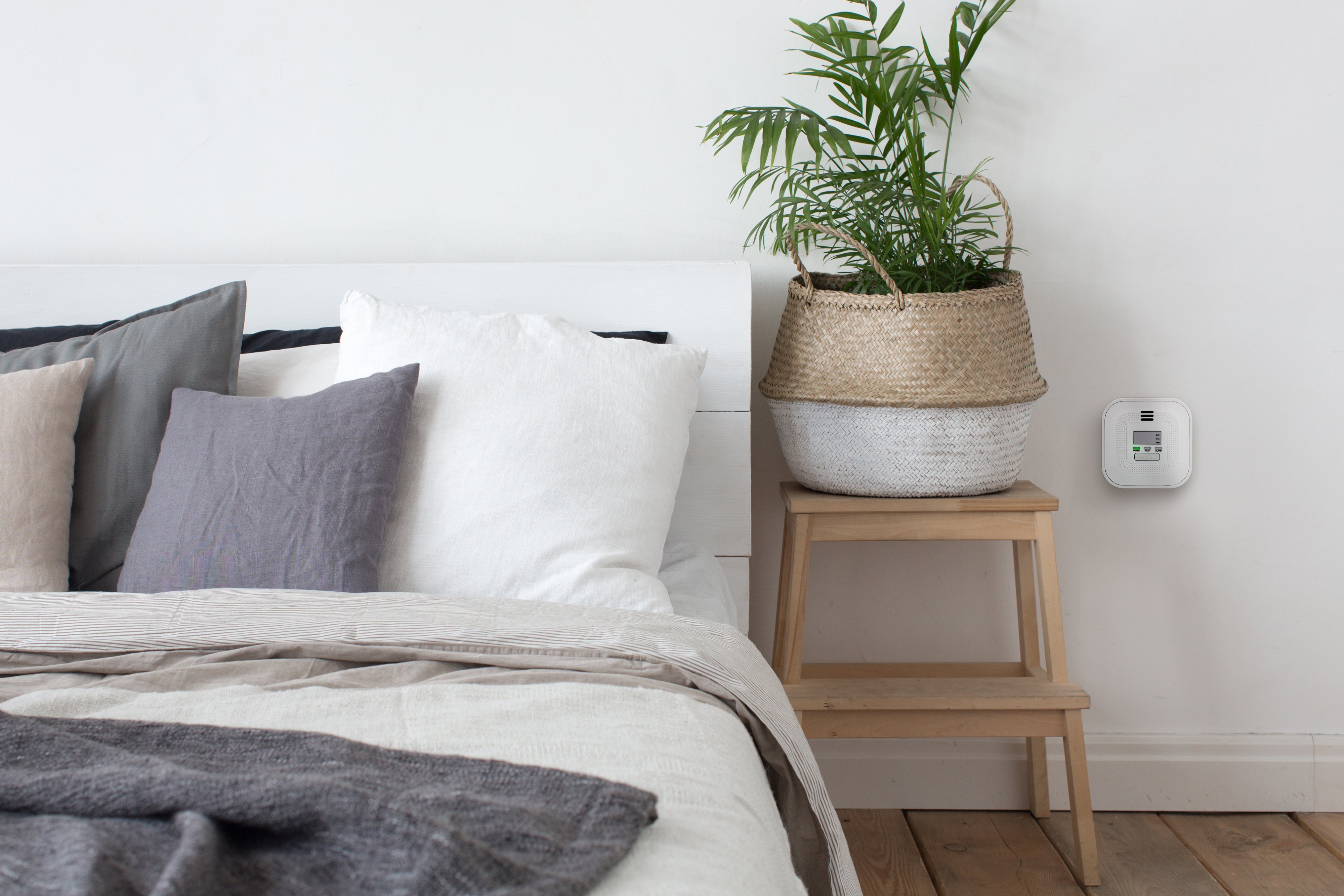 Ein Schlafzimmer mit weißer Wand, Bett mit Kissen, Pflanze im Korb und Melder.