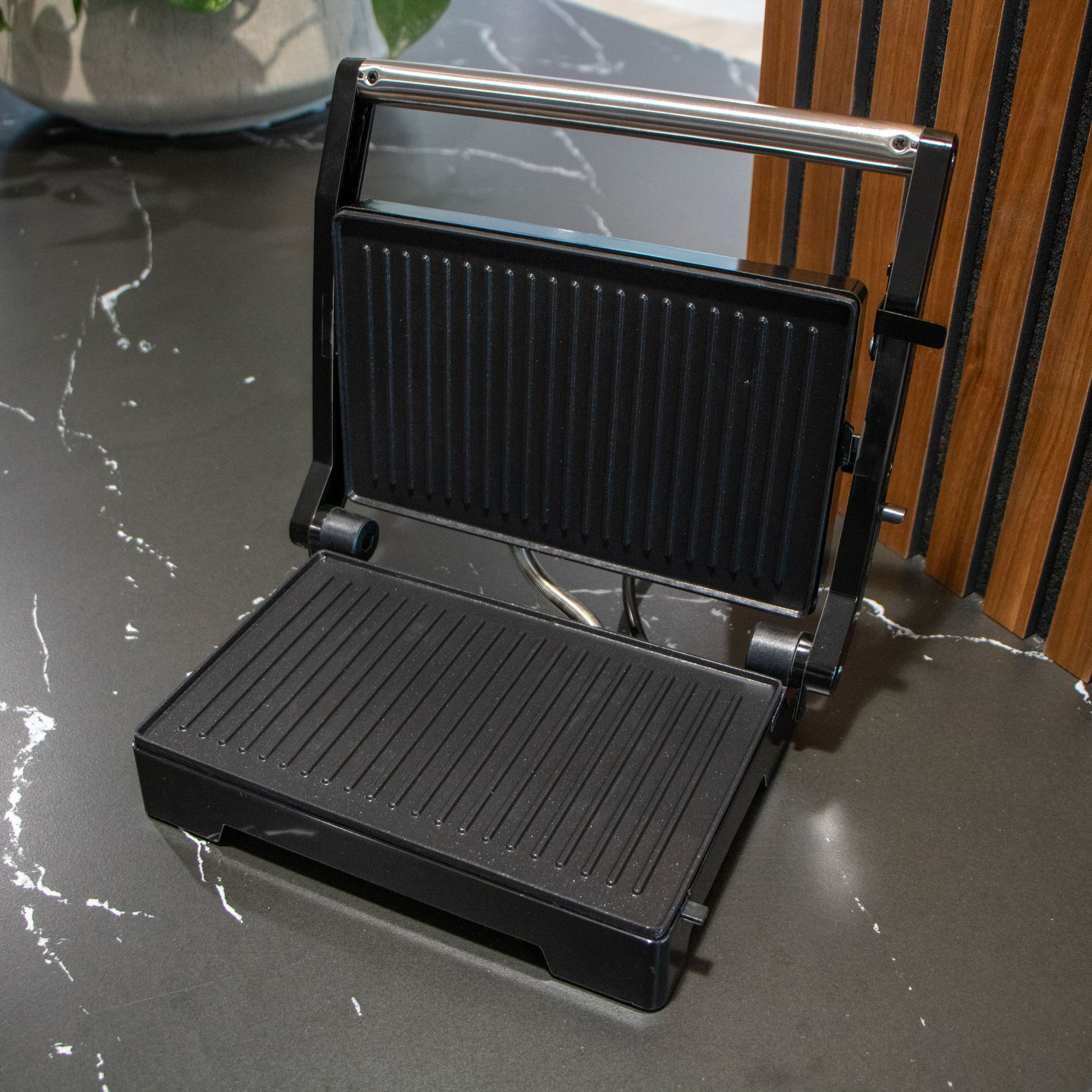 Otwarty czarny grill kontaktowy z żebrowanymi płytami na marmurowym blacie.