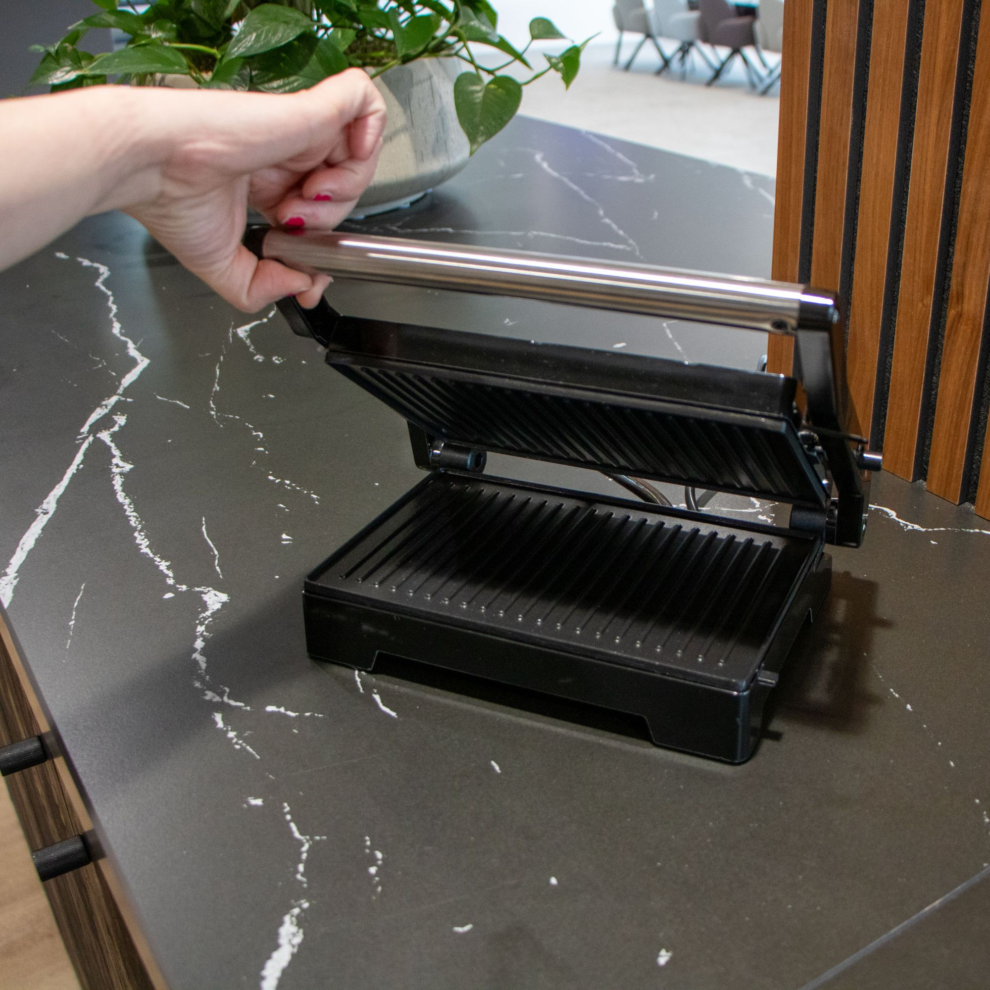 Ręka otwiera czarny grill kontaktowy na ciemnym blacie z wzorem marmuru.
