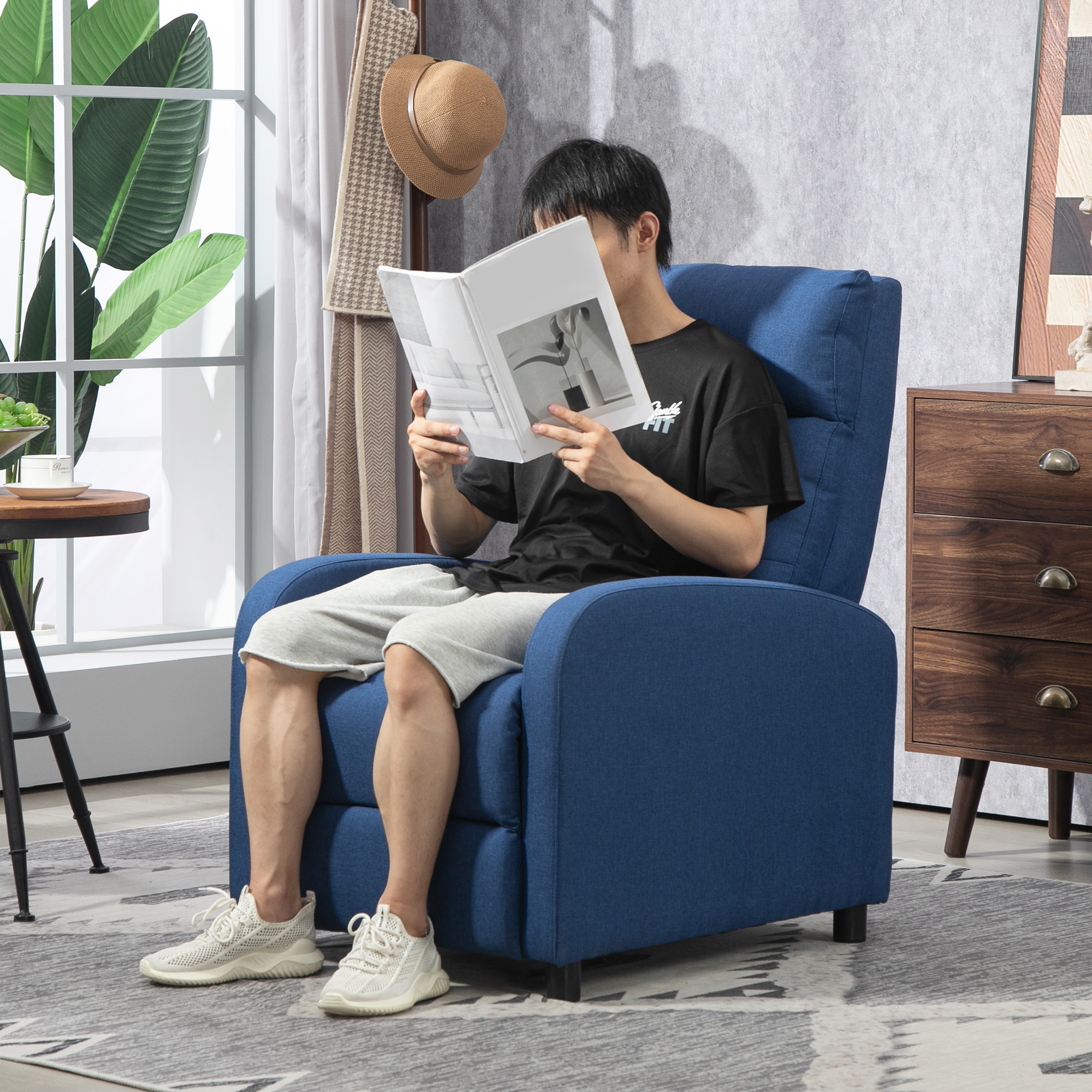 Un hombre lee un libro sentado en un sillón azul en una habitación luminosa.