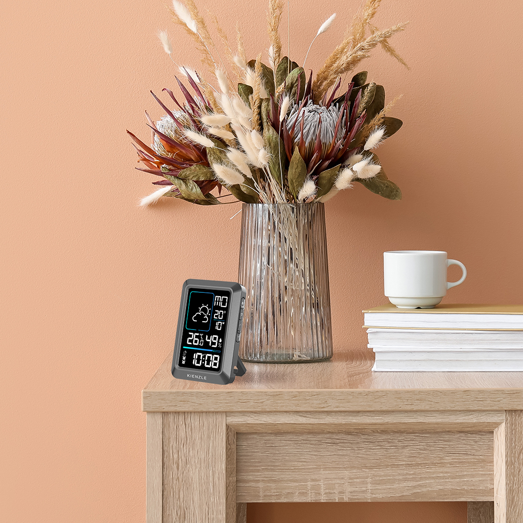 Digitaluhr mit Wetteranzeige steht auf einem Holztisch neben einer Vase mit Trockenblumen.
