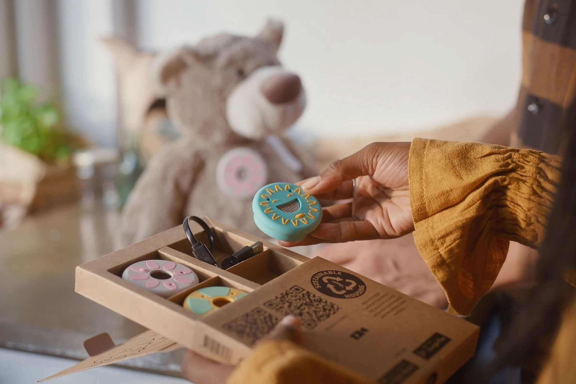 Hand holds blue smiley face charger over gift box with donut charms. Teddy bear behind.