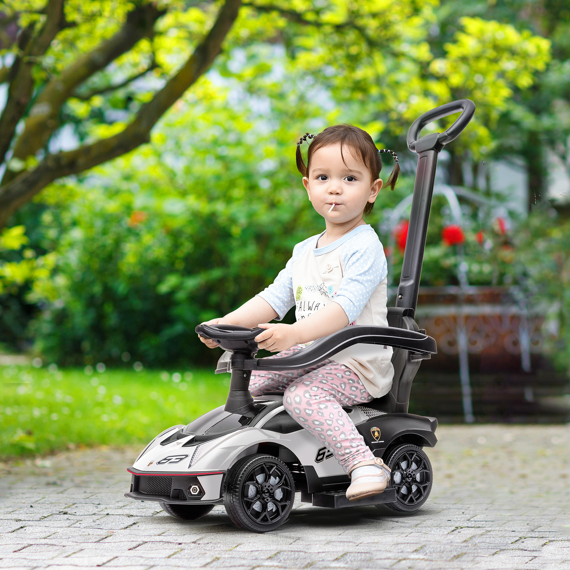 Una niña pequeña se sienta en un coche de juguete plateado con asa, en un jardín.
