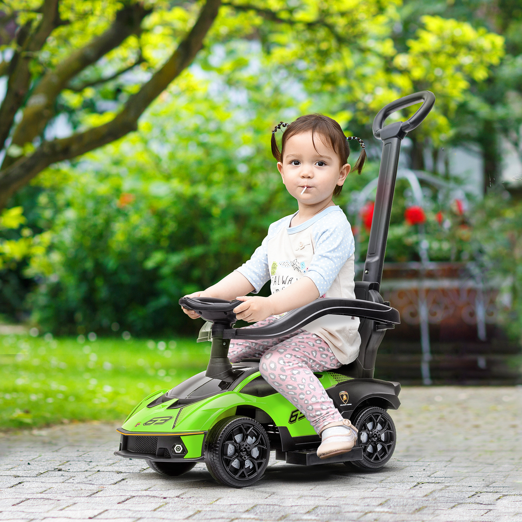 Un niño pequeño se sienta en un coche de juguete verde con asa de empuje, al aire libre en un camino pavimentado.