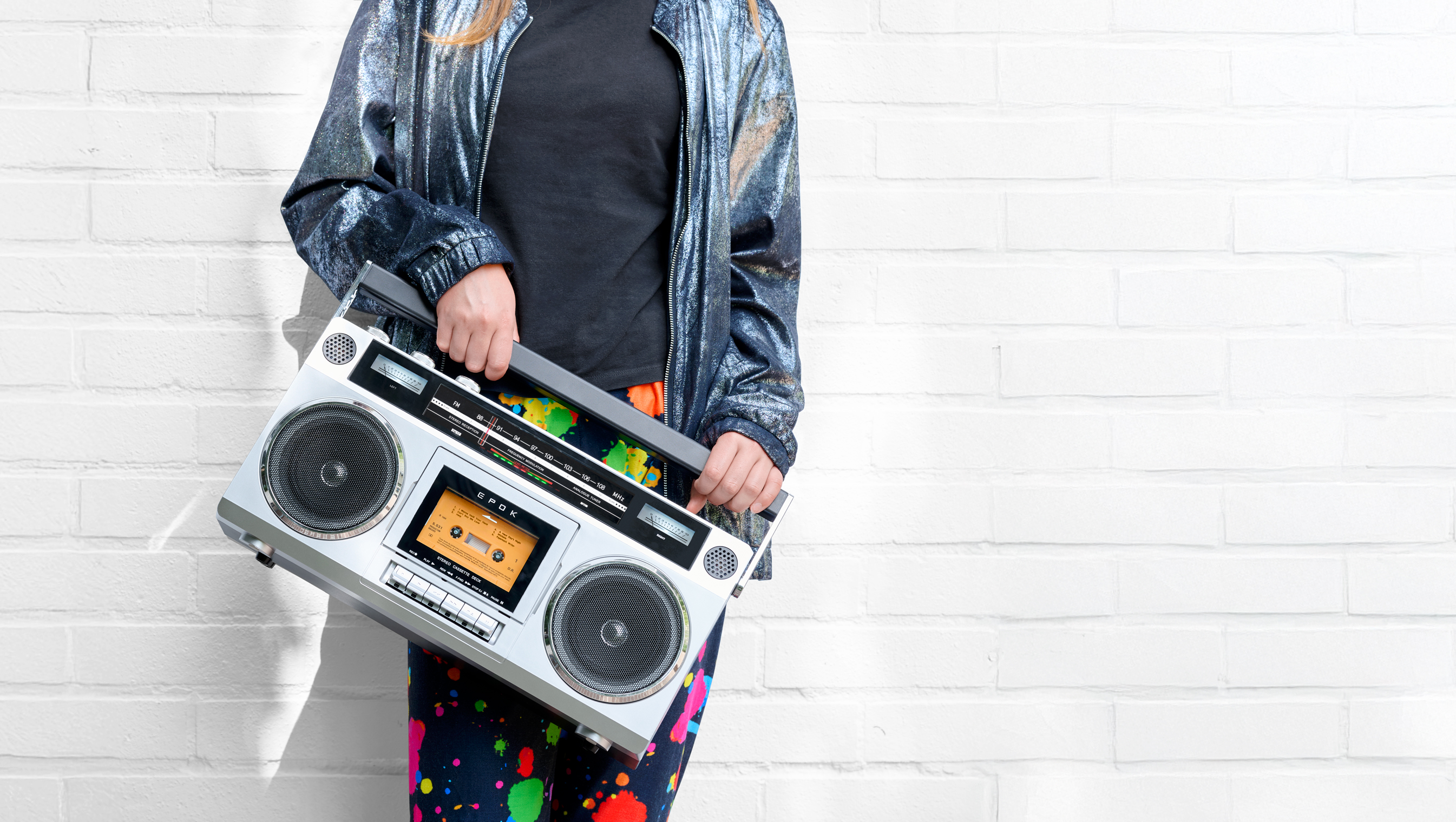 Person hält silbernes Boombox vor weißer Ziegelwand, bunte Hose.