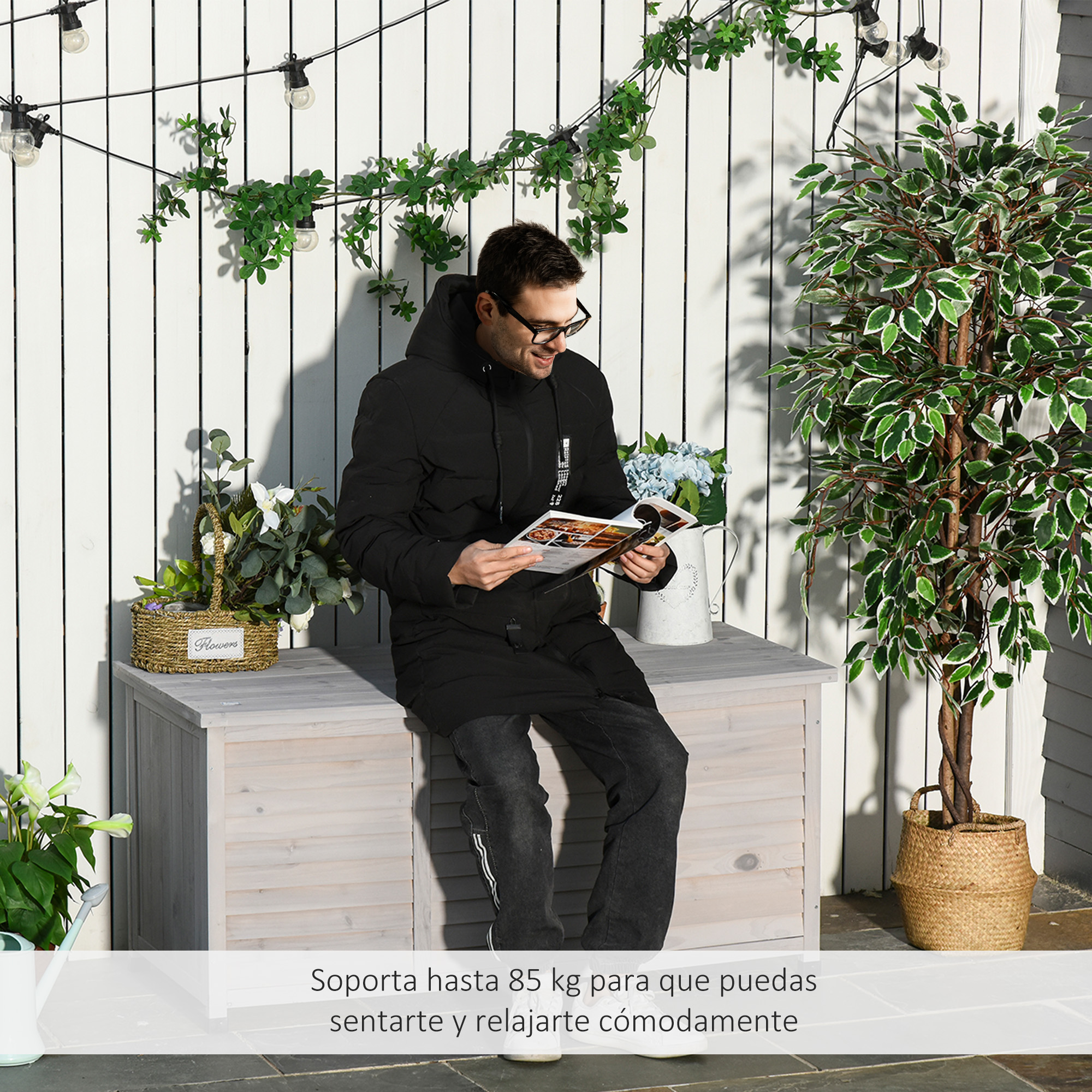 Hombre sentado en cofre de madera, leyendo revista, cerca de planta.
