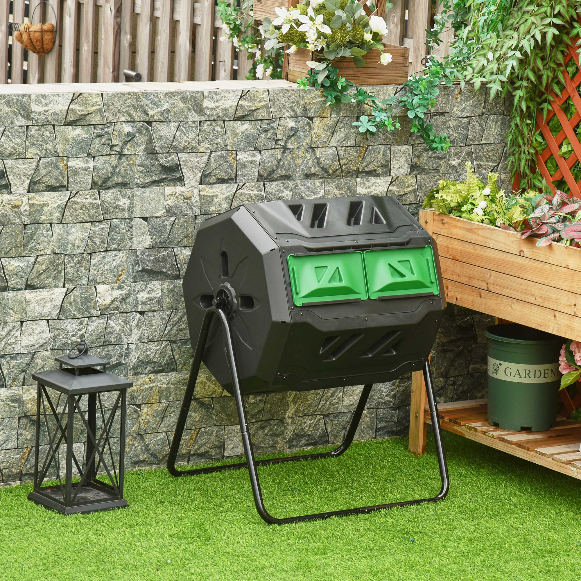 Compostador negro sobre césped artificial verde frente a una pared de piedra.