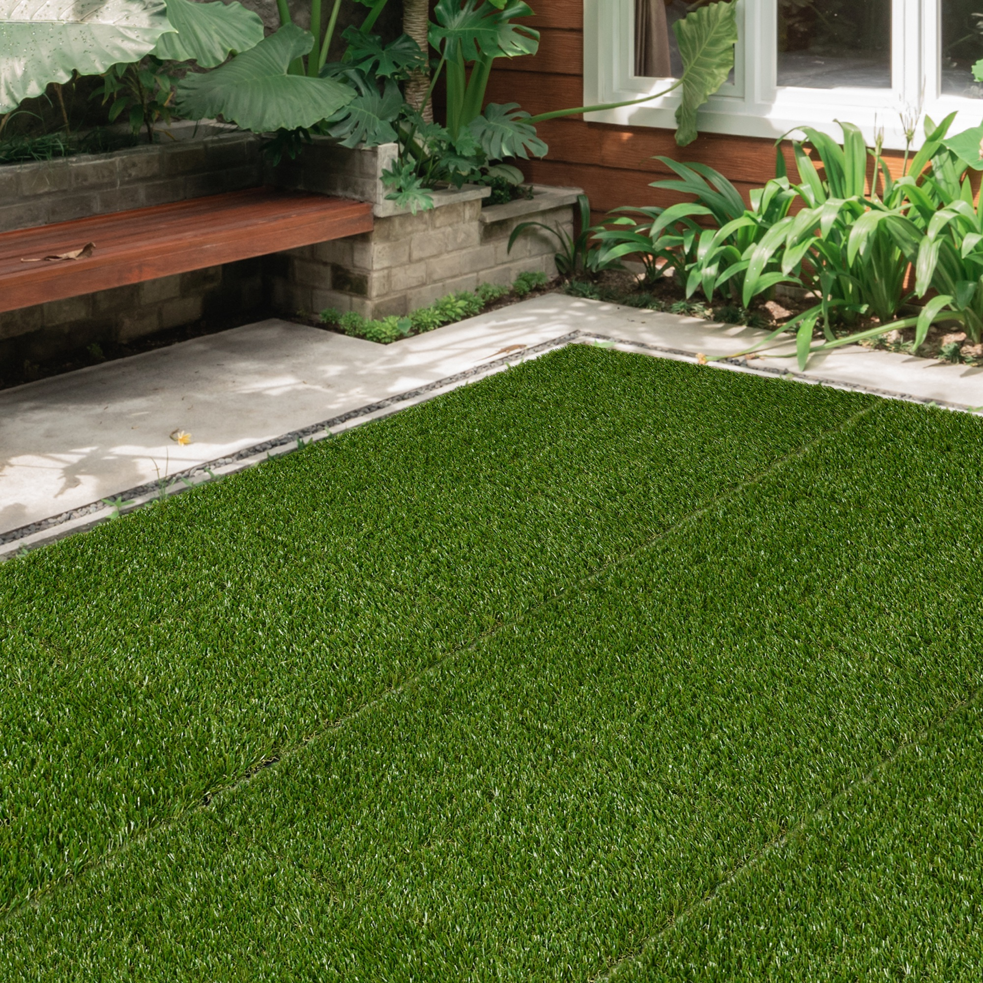 Un área de césped artificial verde bordea un camino de concreto con banco de madera y plantas.