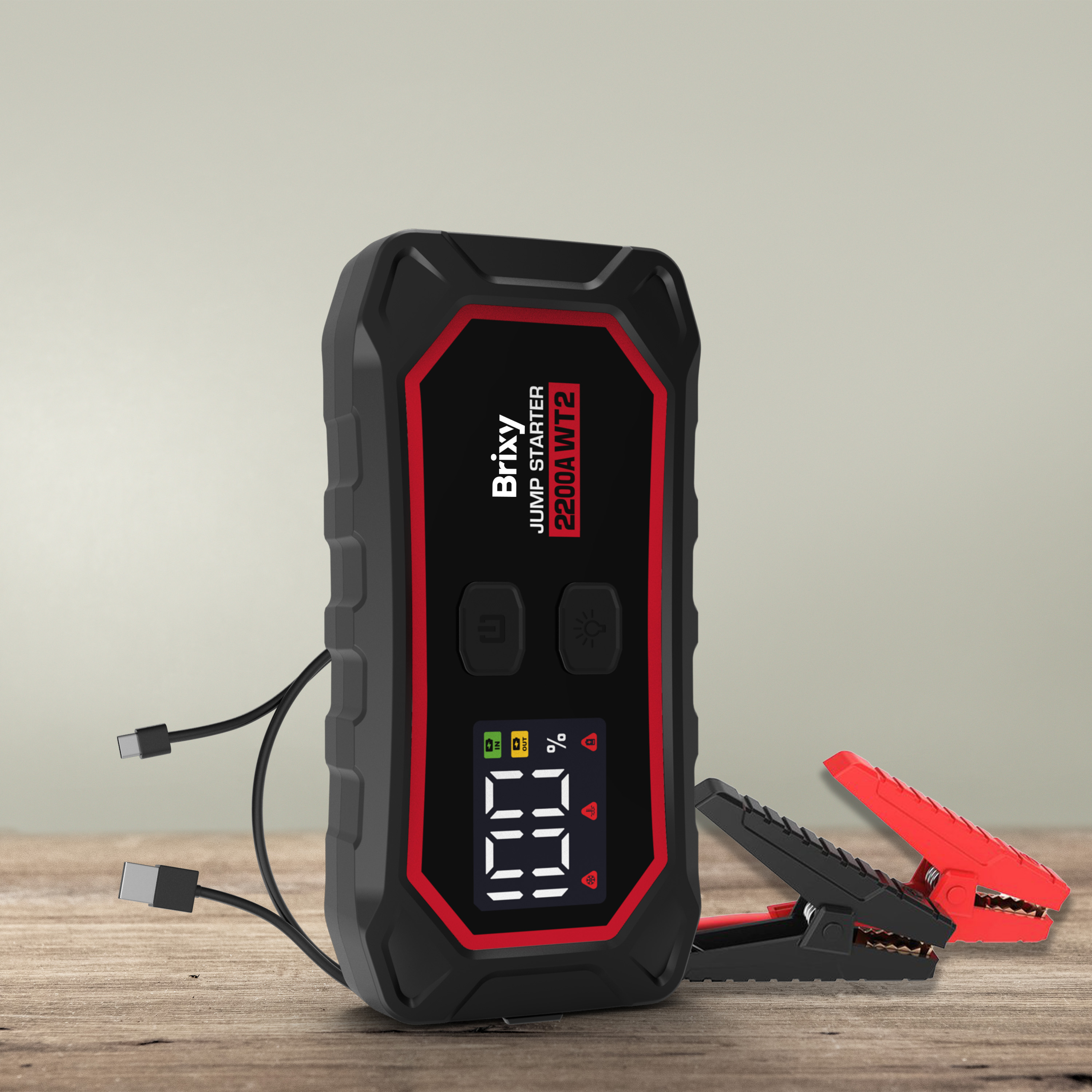 Zwarte startbooster met rode rand, kabels en klemmen op hout.
