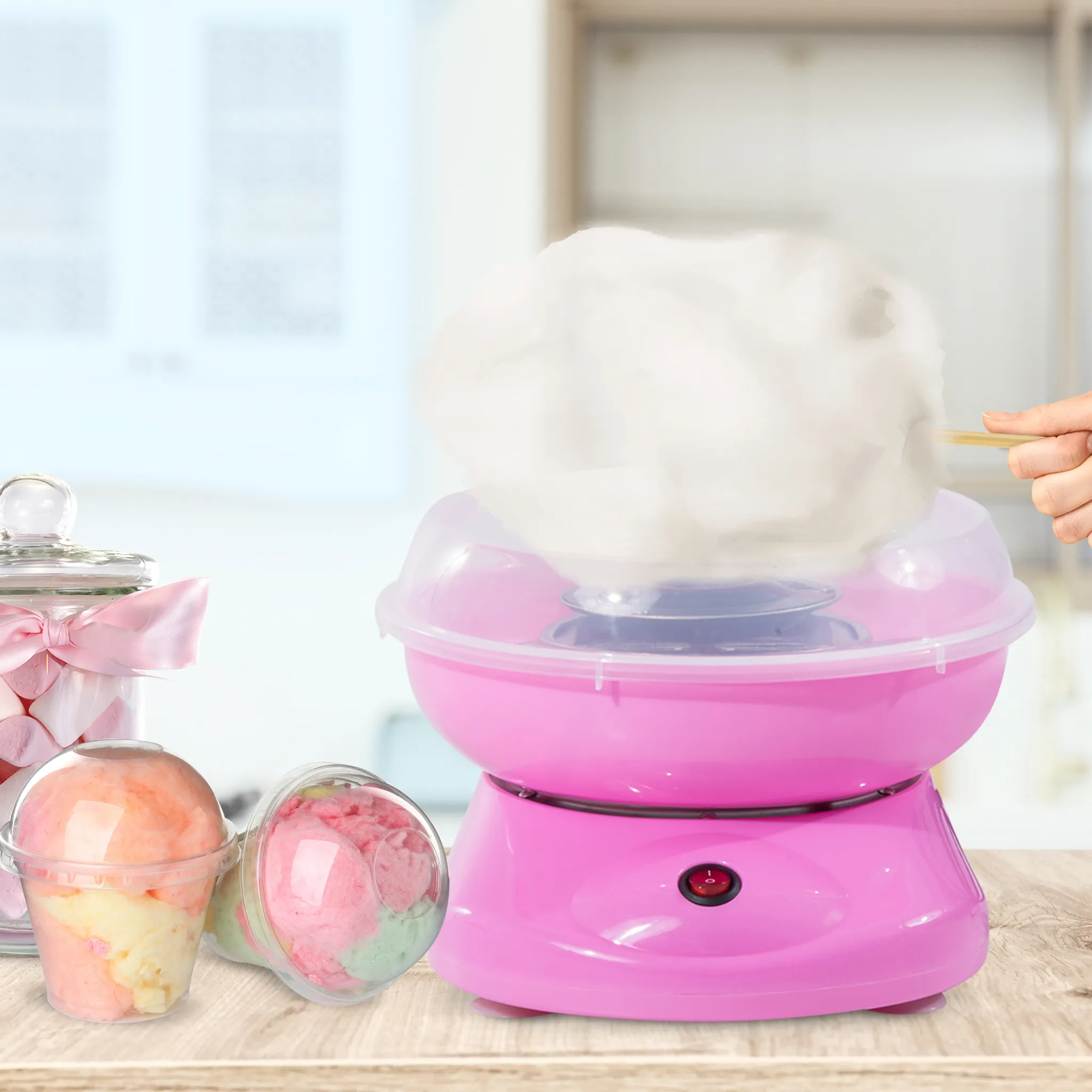Eine rosafarbene Zuckerwatte-Maschine mit weißer Zuckerwatte, daneben Becher mit Zuckerwatte und ein Glas Marshmallows.