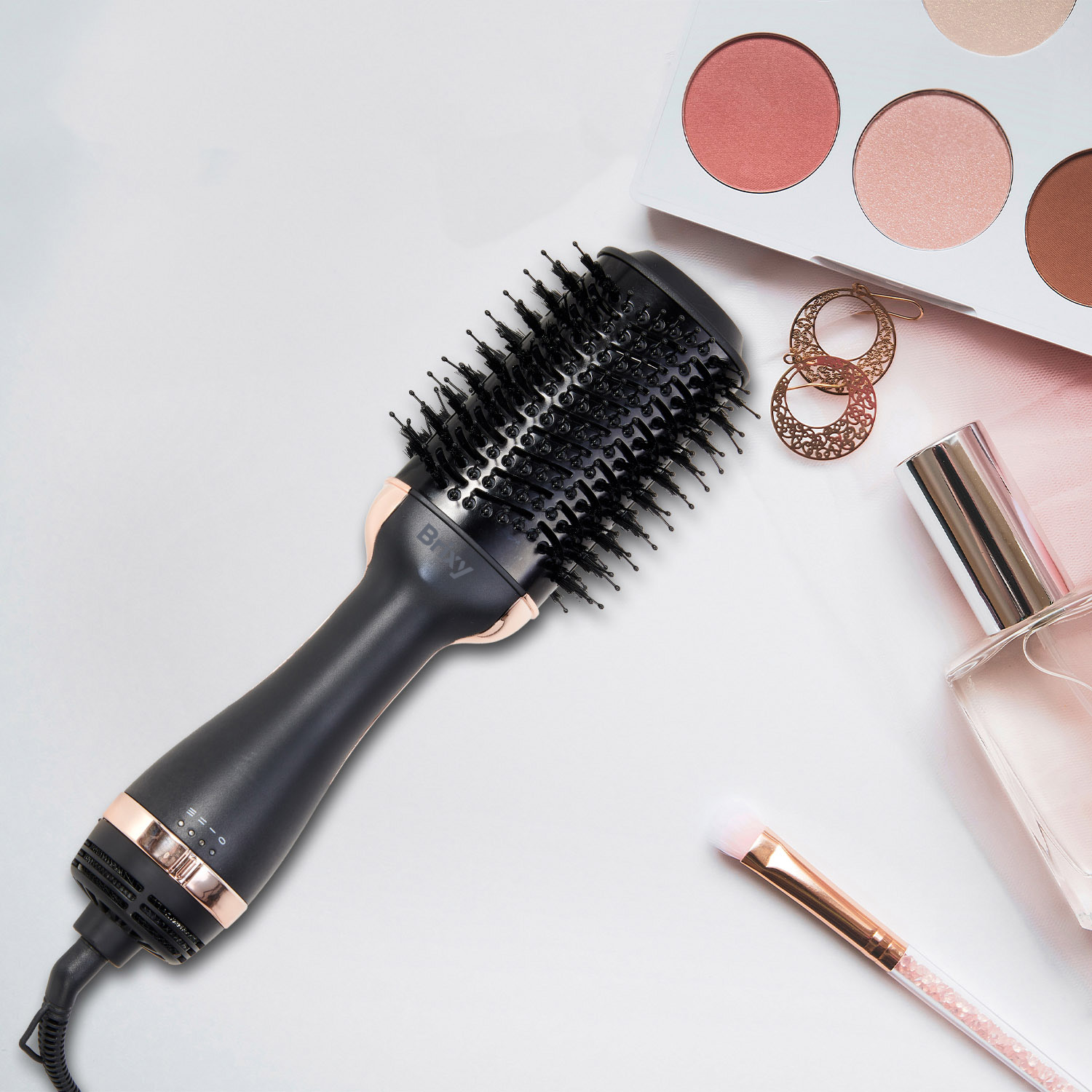 Een zwarte haardroger met roségouden accenten, make-uppalet, oorbellen, parfum en kwast.