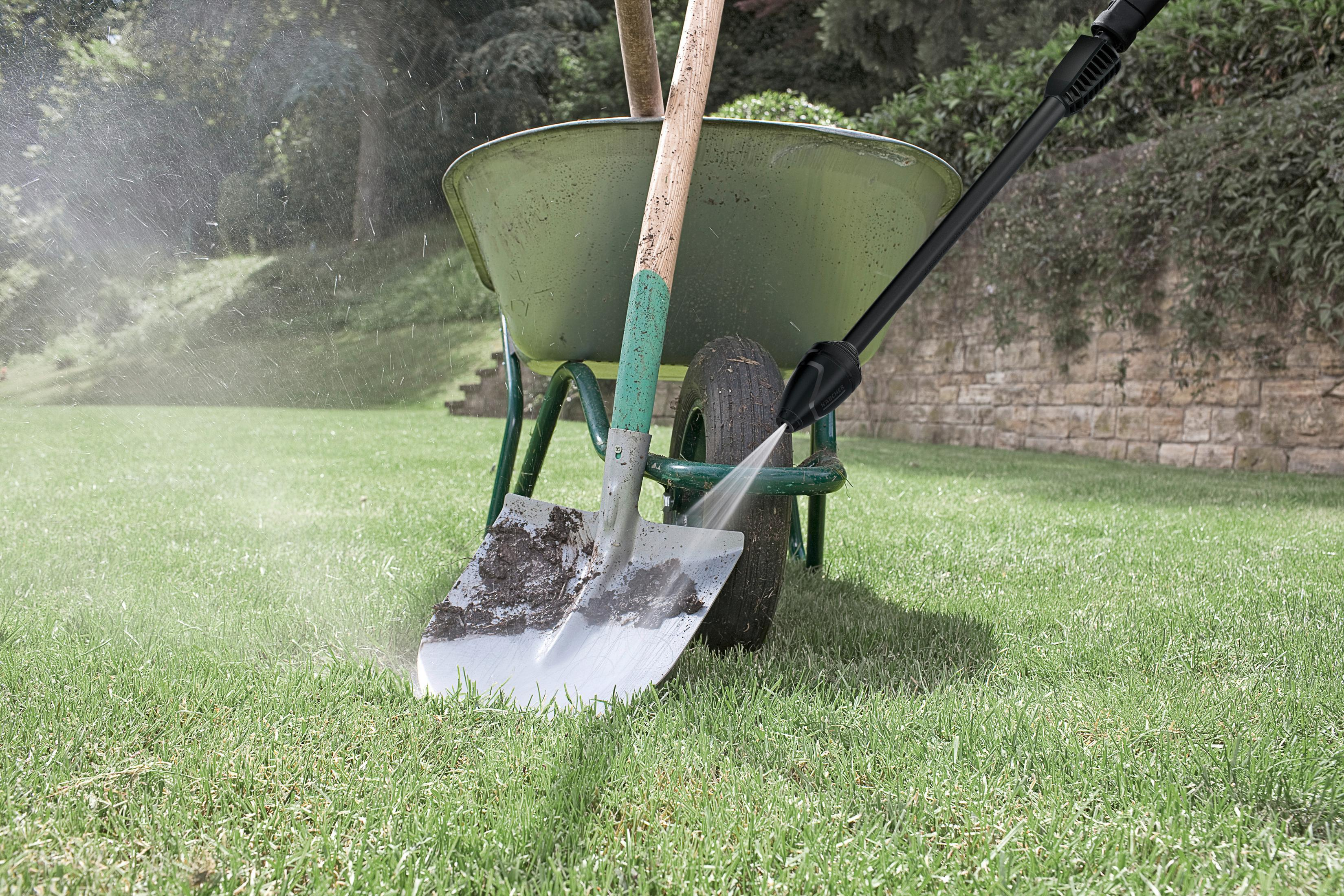 A pressure washer cleans a dirty shovel next to a green wheelbarrow on grass.