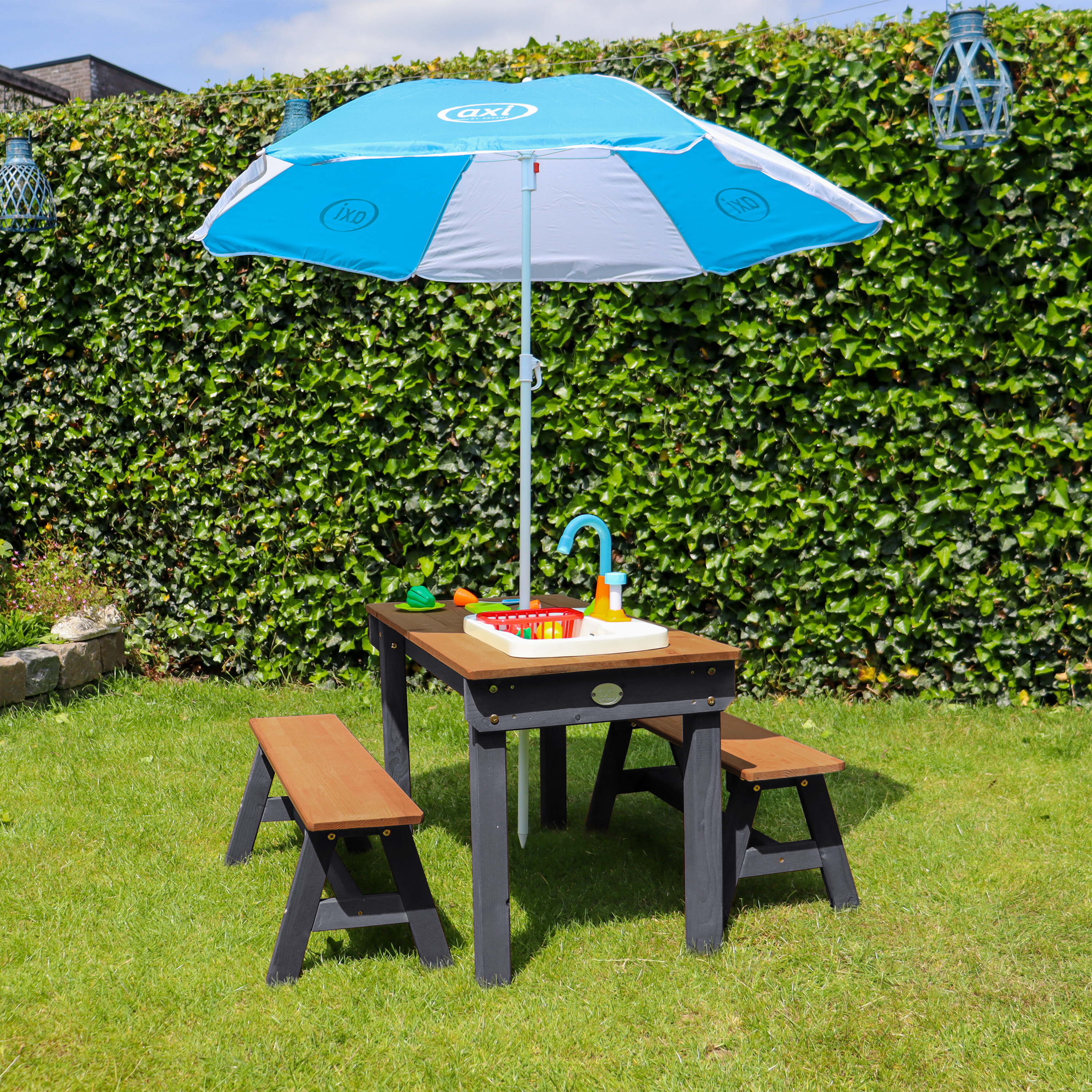 Ein Kindertisch mit Spielzeugspüle unter einem Sonnenschirm im Garten.