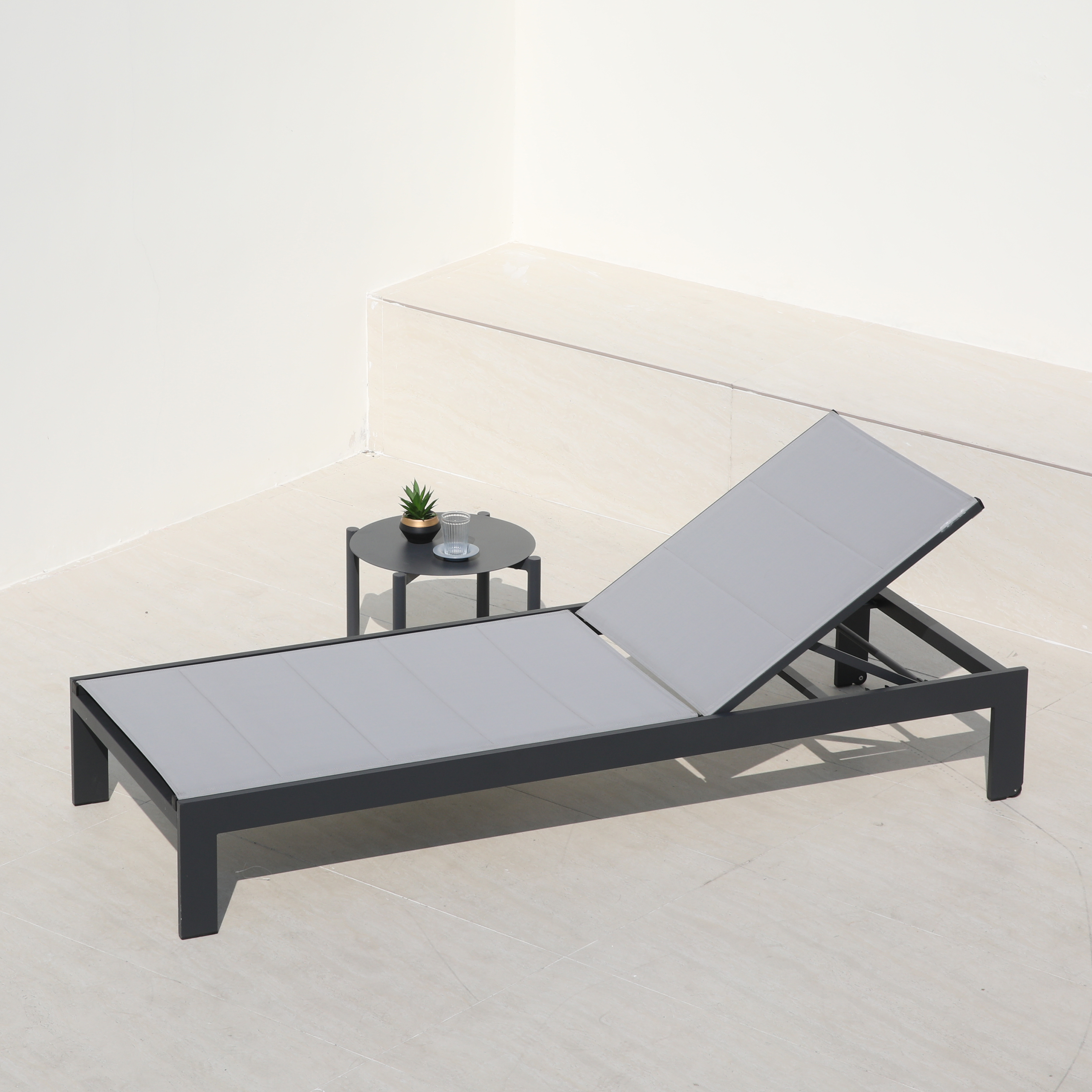 Ein grauer Liegestuhl steht auf einer hellen Terrasse neben einem kleinen Tisch mit Pflanze und Glas.