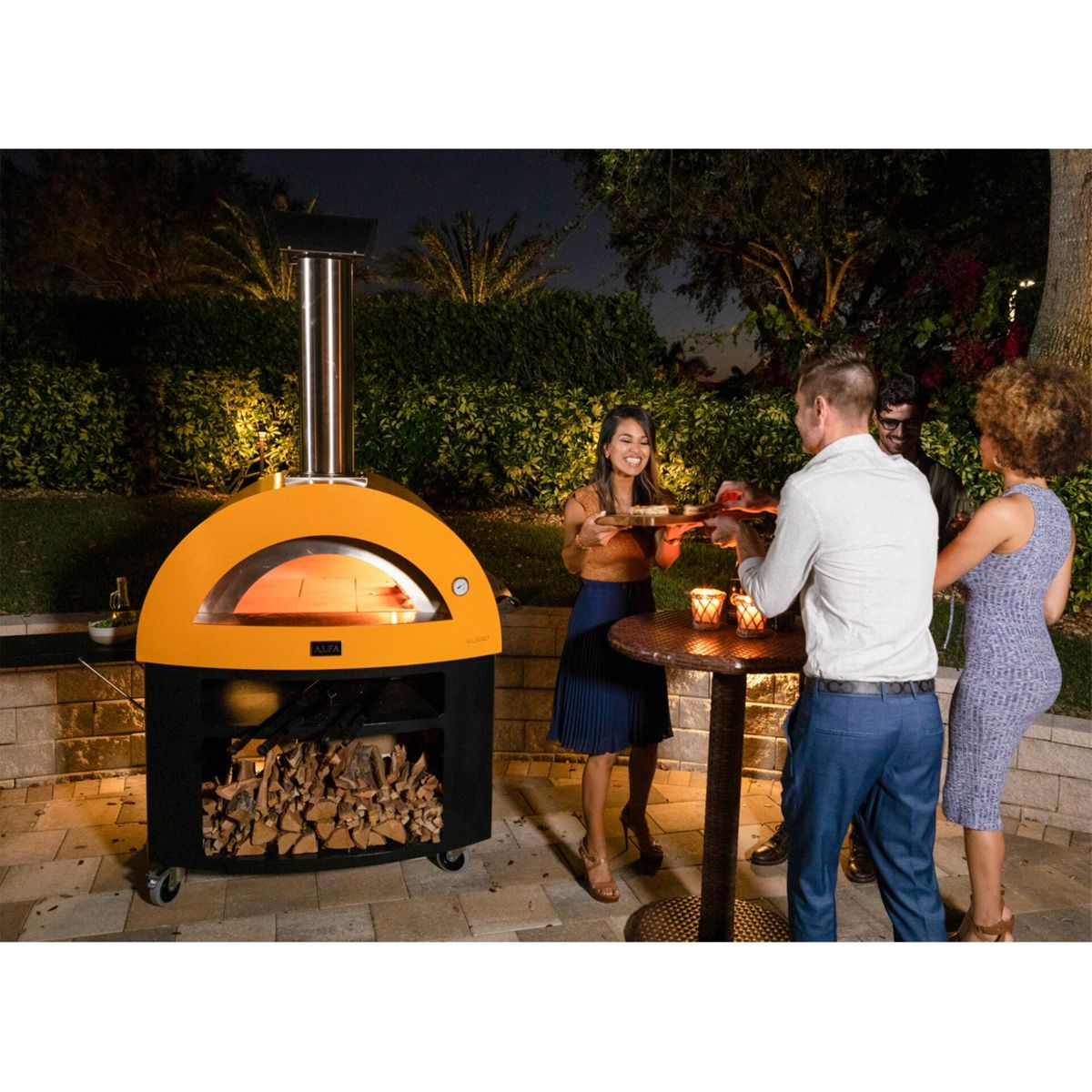 Menschen feiern abends um einen Pizzaofen und einen Tisch mit Kerzen.