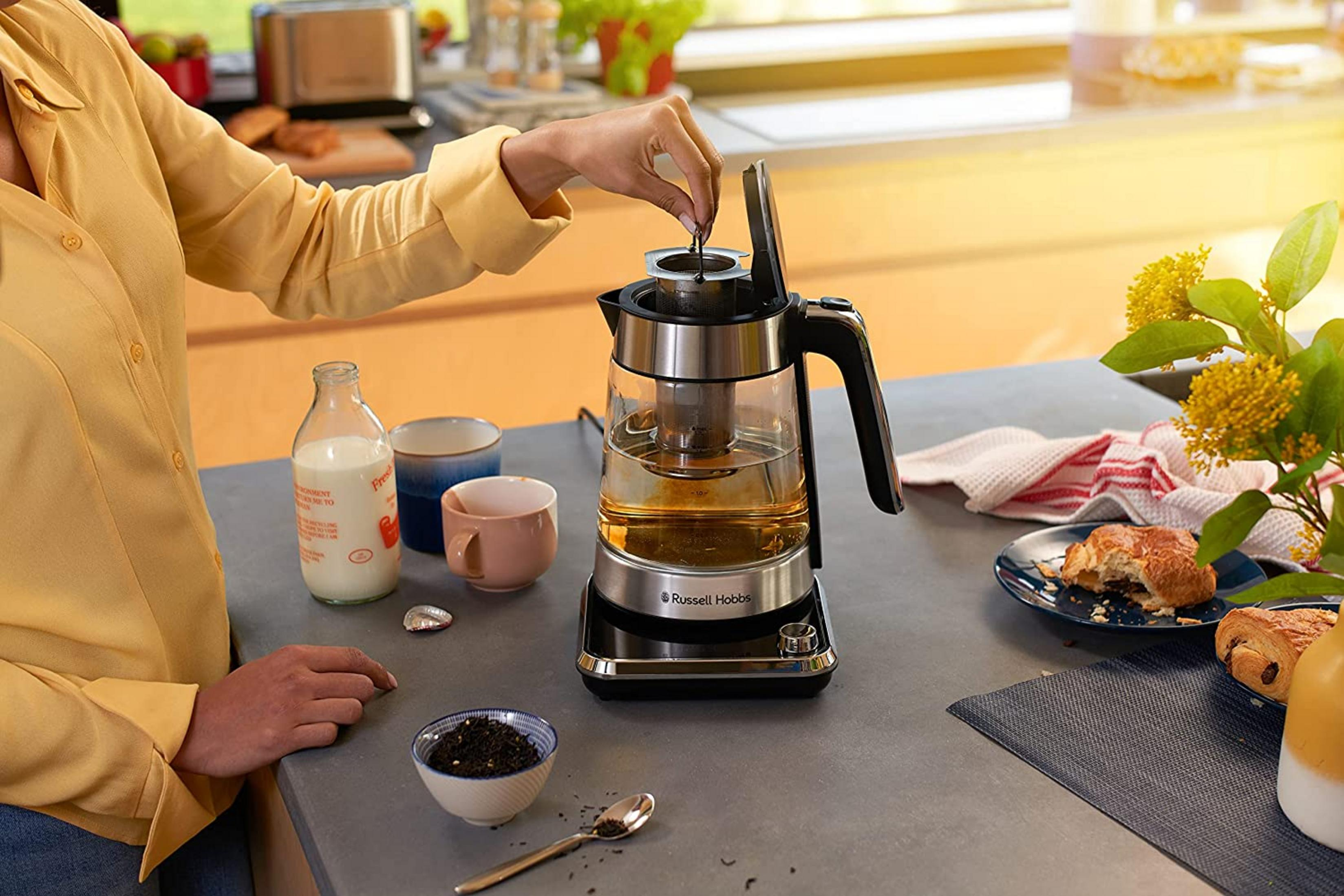 Une femme prépare du thé. Une bouilloire, du lait, des tasses et des pâtisseries sont sur la table.
