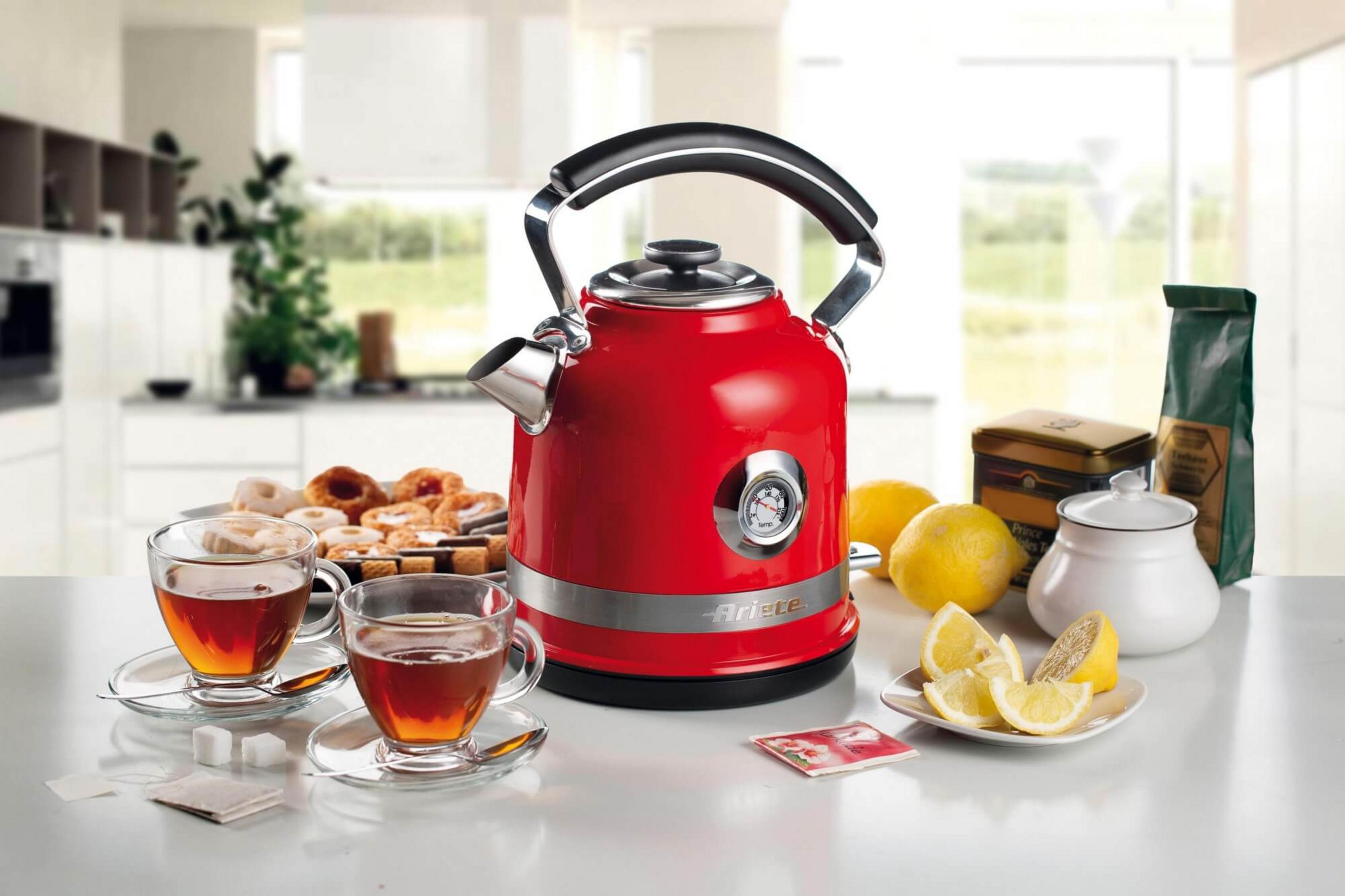 Een rode waterkoker met thermometer, kopjes thee, gebak, citroenen en theezakjes.