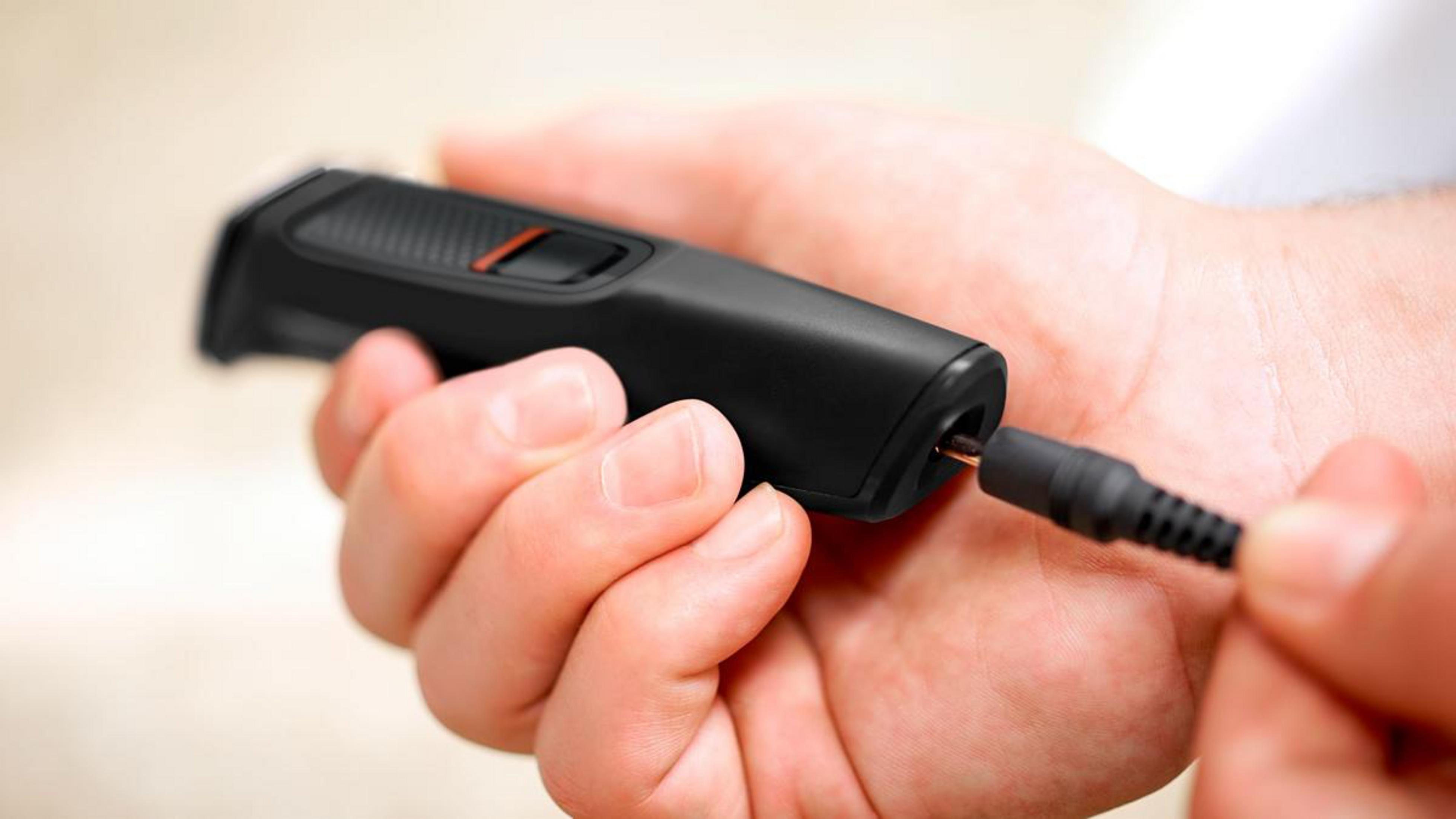 A hand holds a black trimmer as a charging cable is plugged in.