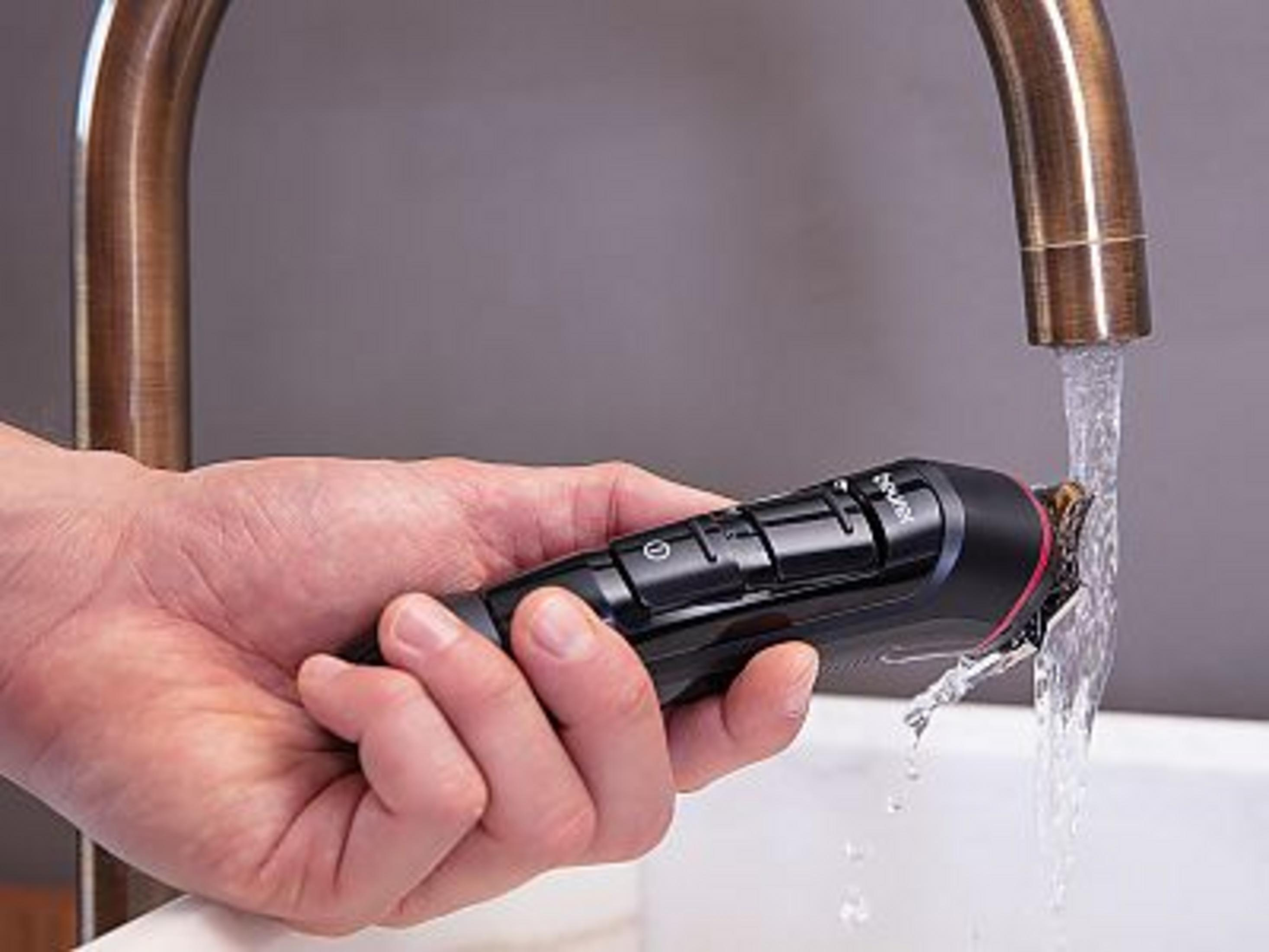 Een hand houdt een zwarte trimmer onder stromend water in een wastafel.