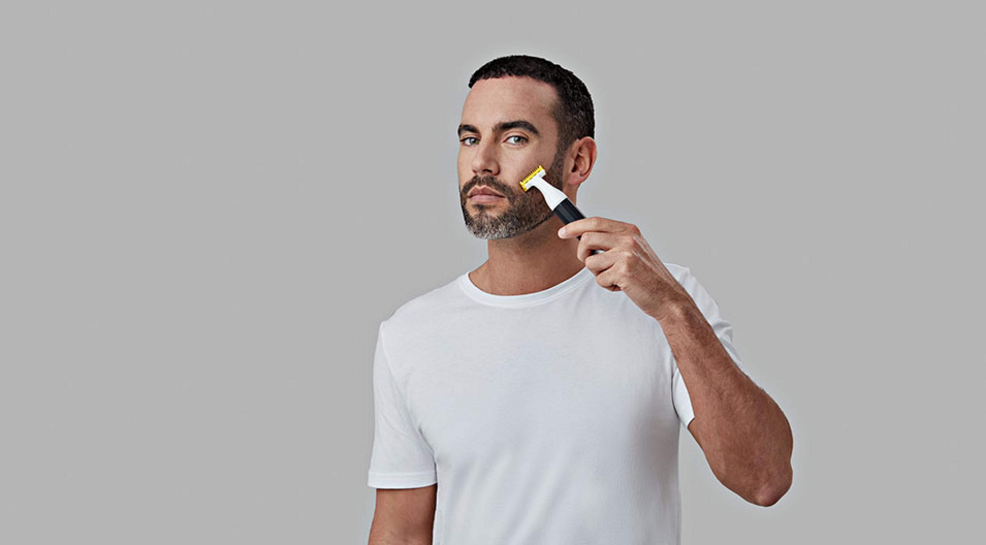 Mann mit Bart rasiert sich im Gesicht mit einem Trimmer auf grauem Hintergrund.