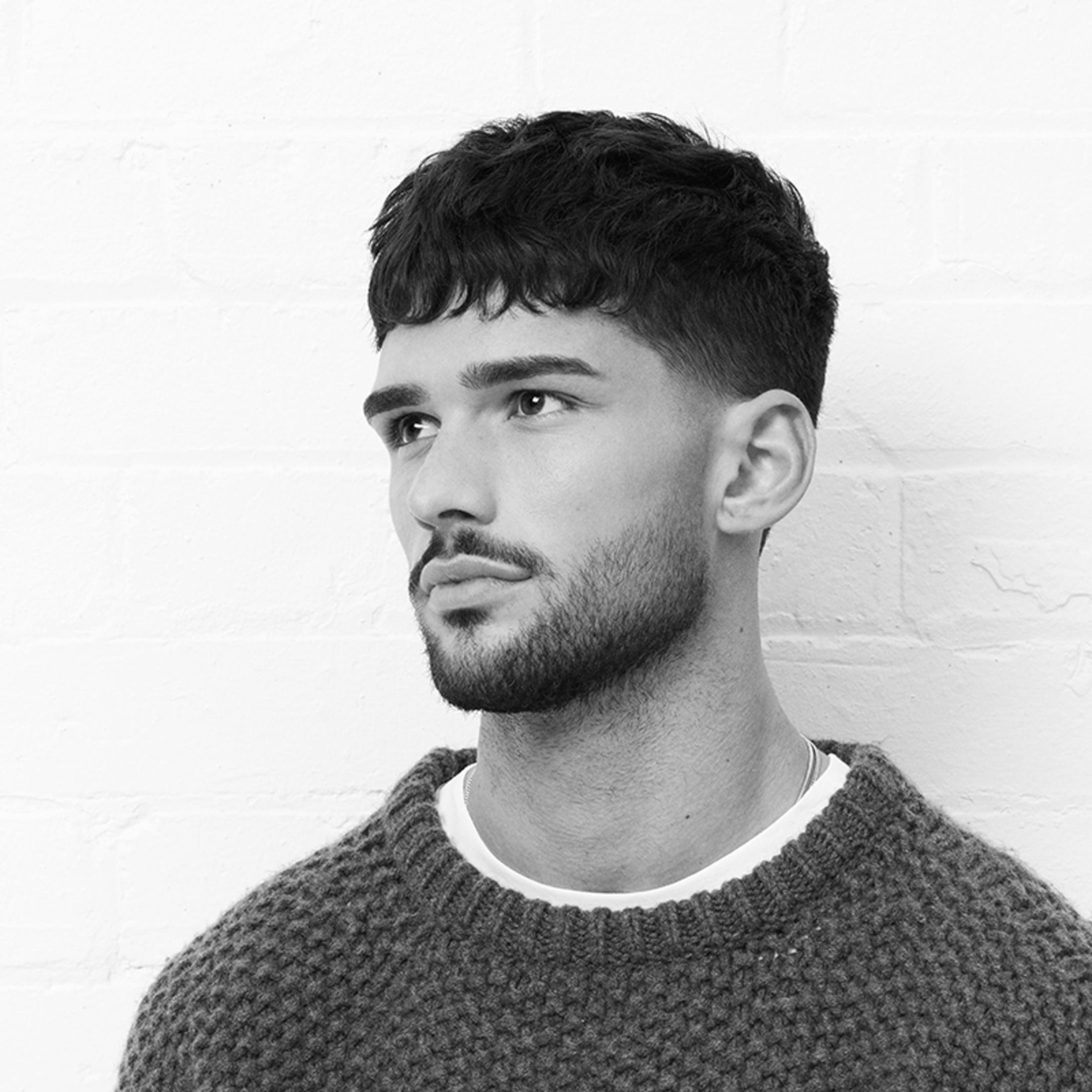 Mężczyzna z krótkimi, falowanymi włosami i brodą nosi sweter.