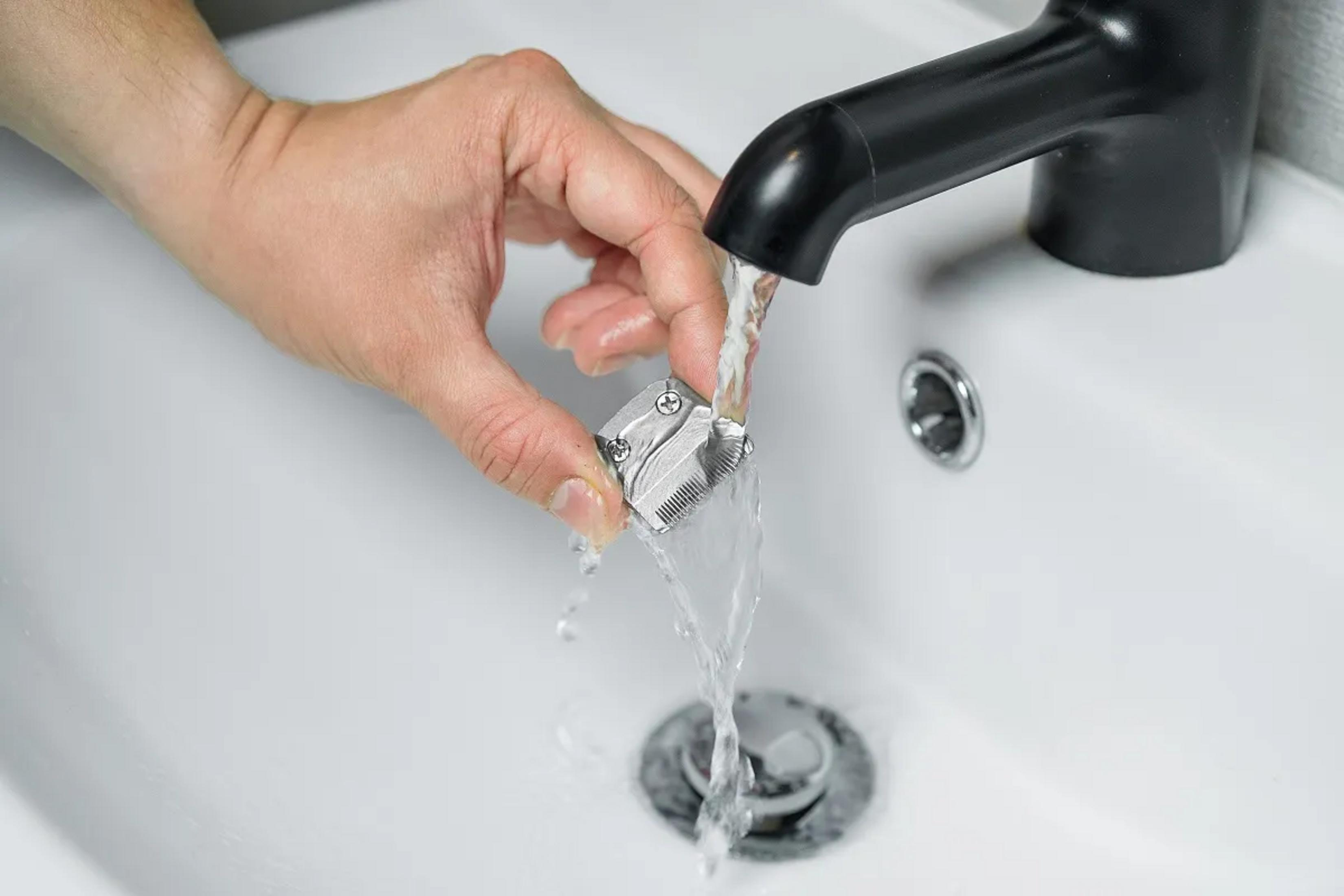 Dłoń płucze srebrną głowicę trymera pod bieżącą wodą w białej umywalce.
